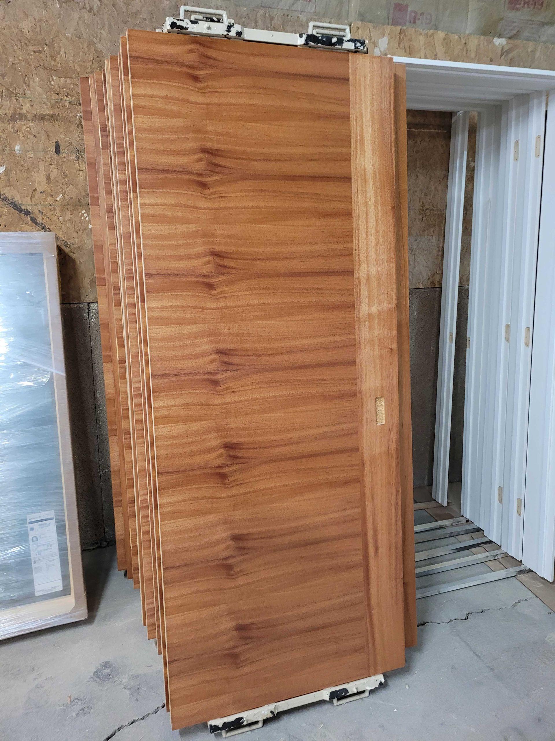 A stack of horizontal-grain wooden sliding doors with metal hardware, leaning in a workshop near white door frames.