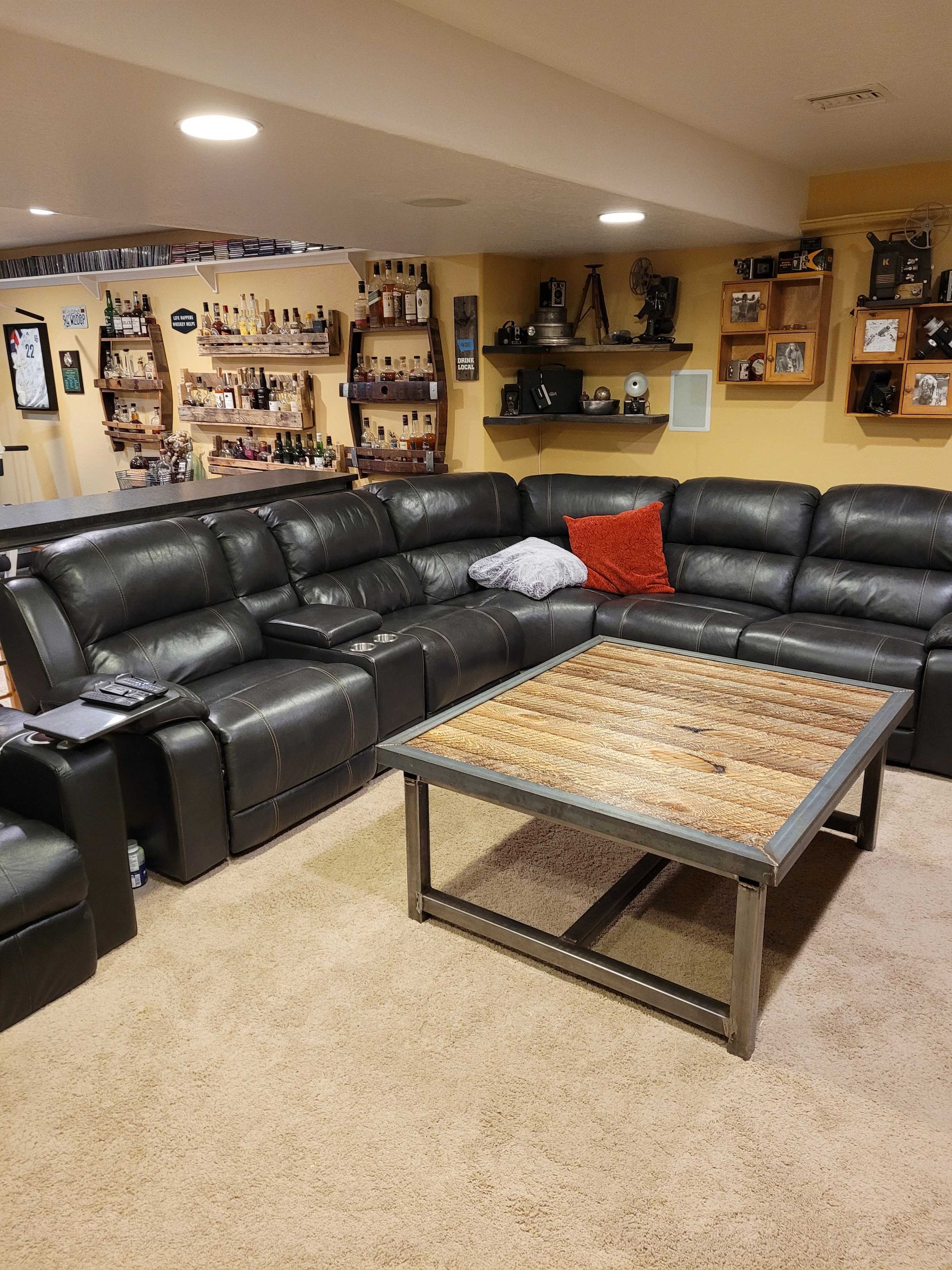 A large black leather sectional sofa with a wooden industrial coffee table in a finished basement with shelves of decor.