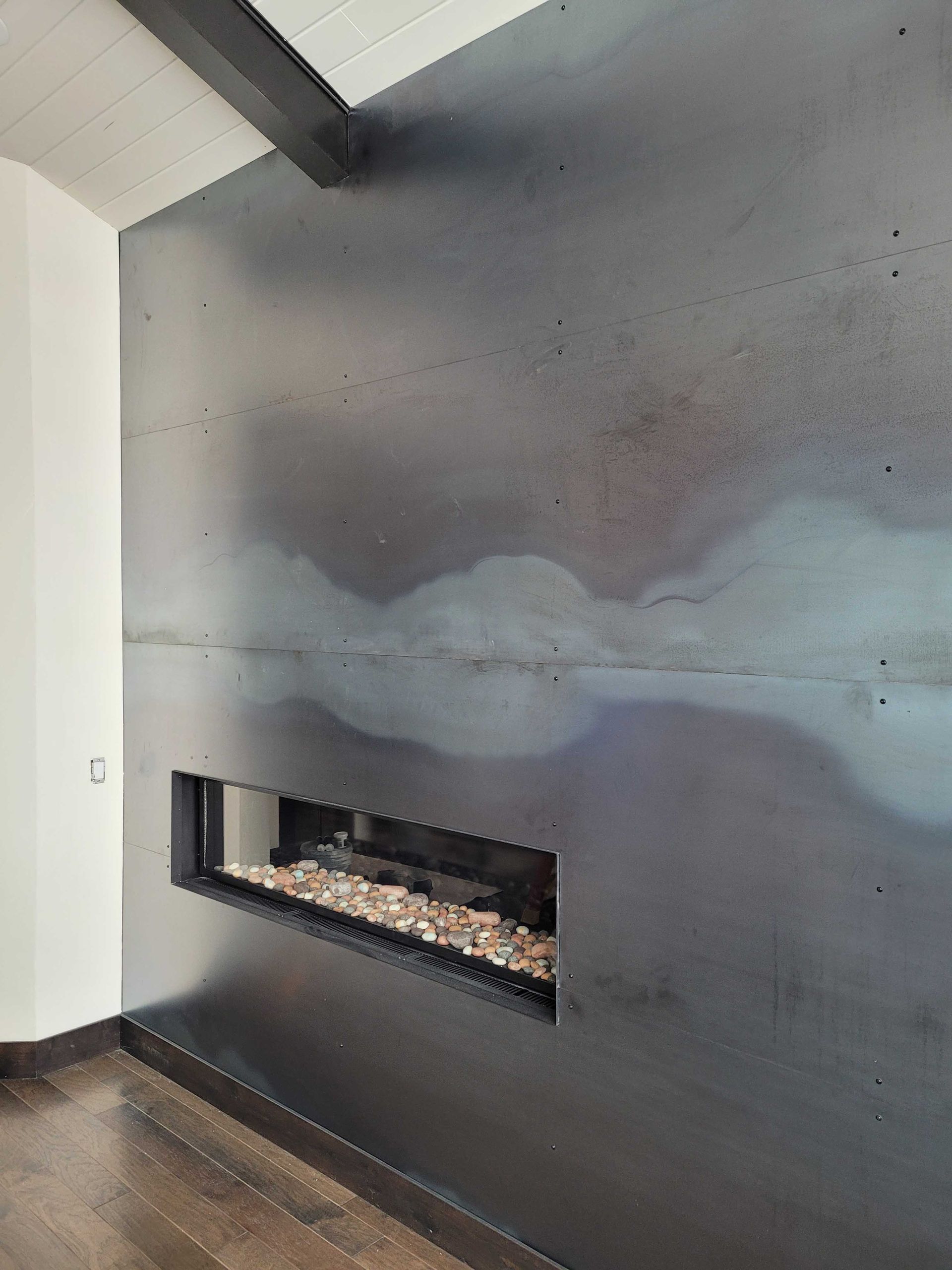 A contemporary fireplace wall clad in dark, patinated steel plates with a central linear hearth filled with light stones.