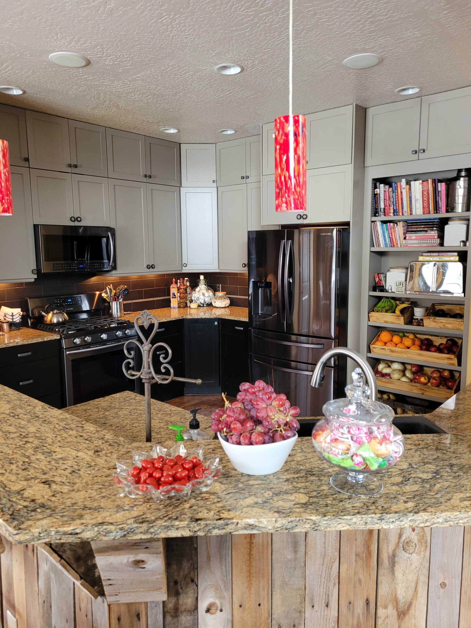 A kitchen with grey cabinets, black appliances, a granite countertop with fruit, and an open shelving pantry.