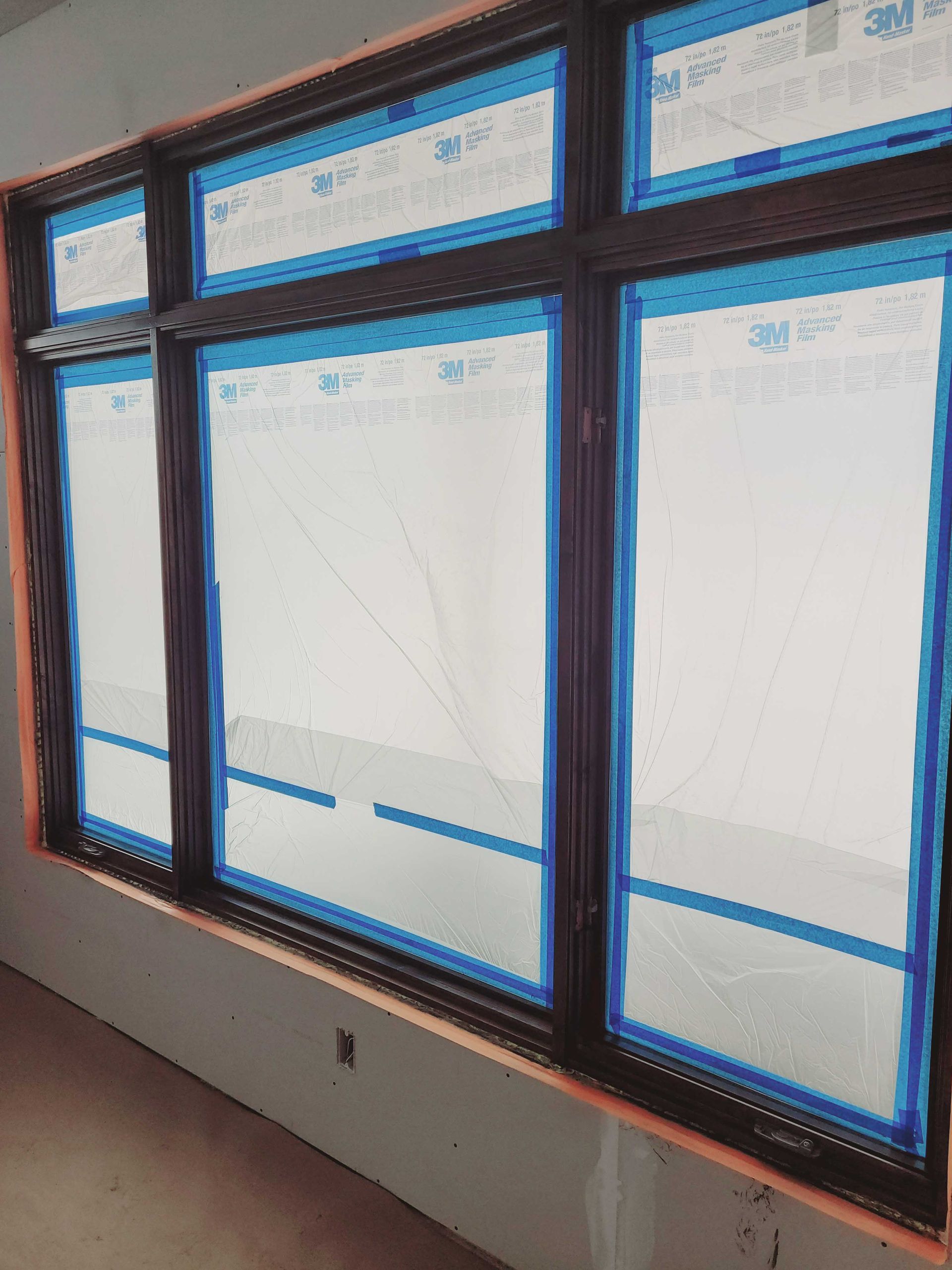 A large, dark-framed window unit in a room under construction, with blue painter's tape covering the glass panes.