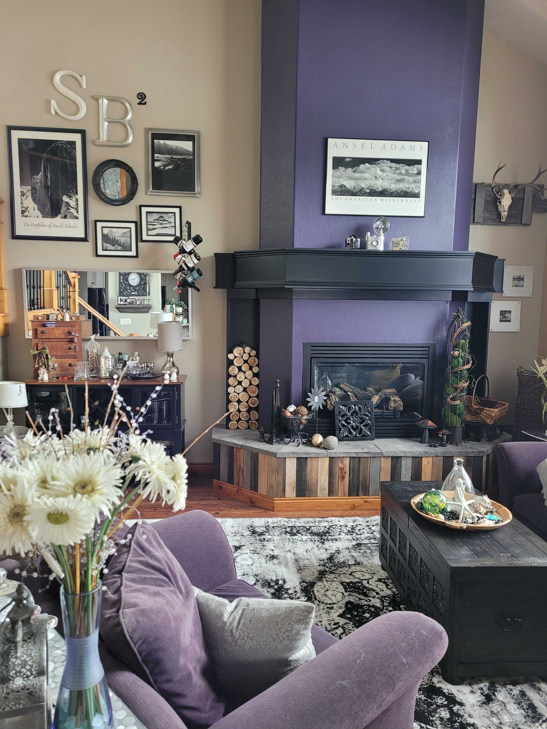 A cozy living room features a purple fireplace, a black mantel, a wood stack, and a gallery wall with S and B letters.