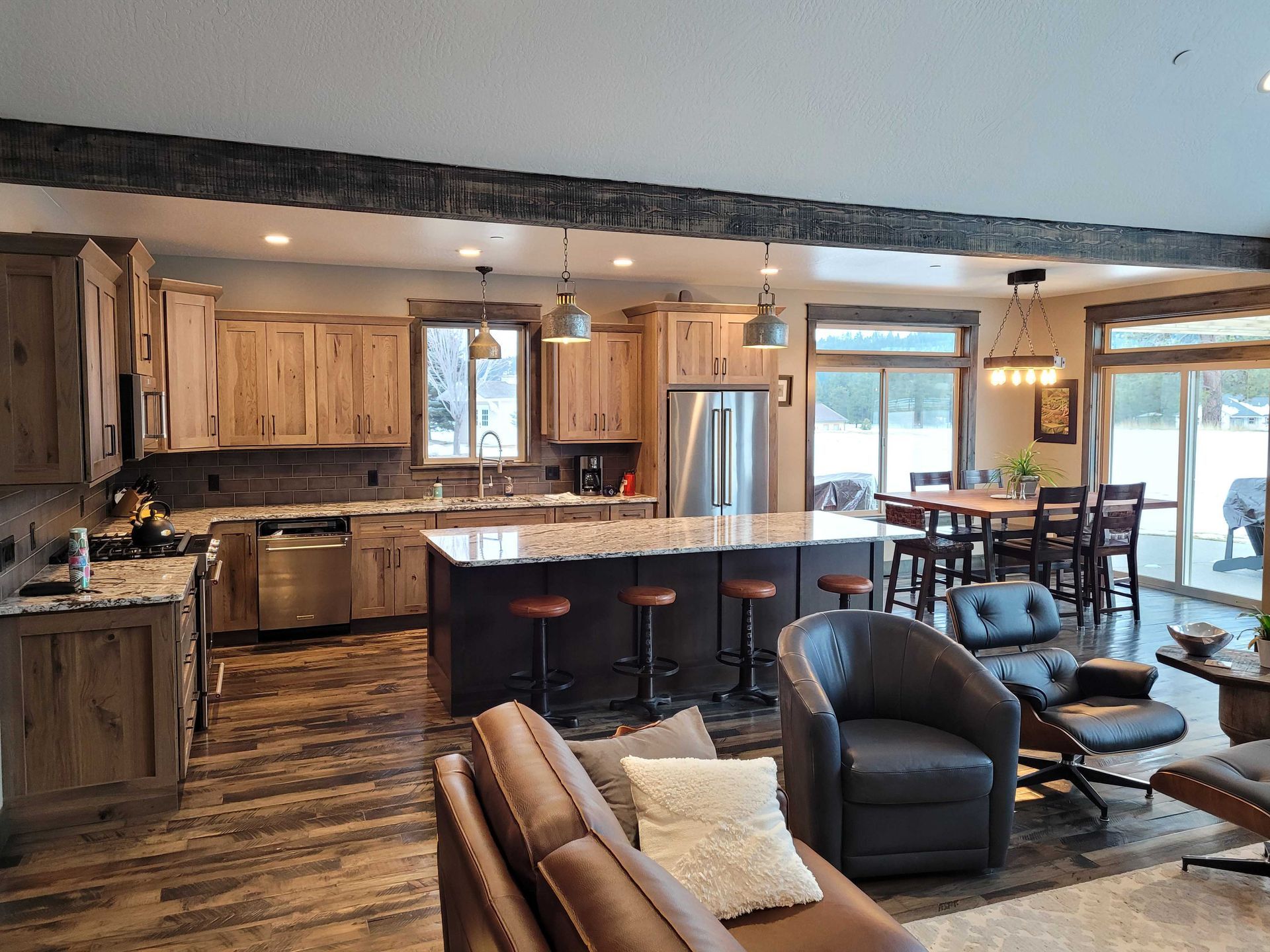 A modern open-plan kitchen and living room featuring light wood cabinets, a granite island with stools, and dark floors.