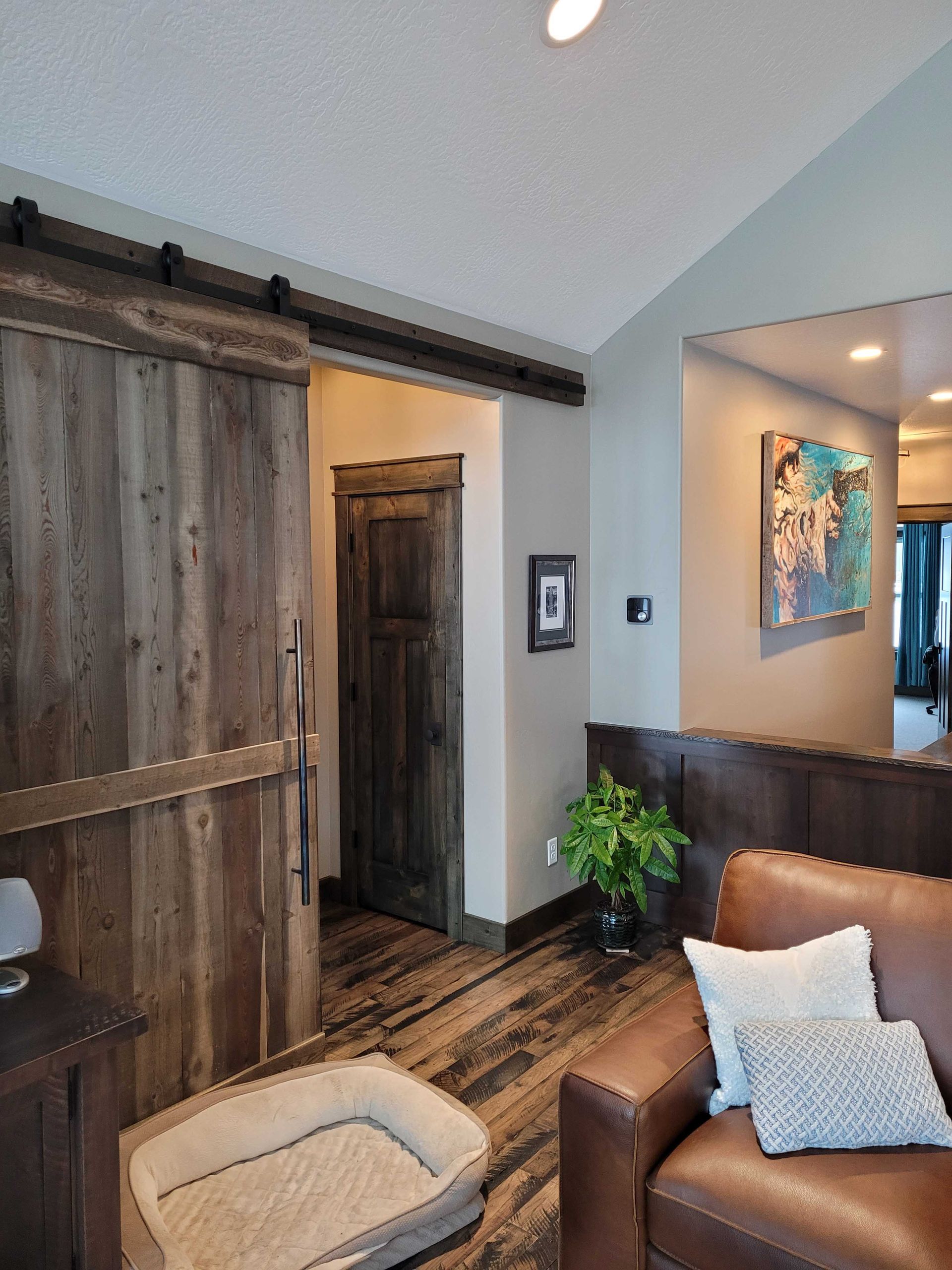 A room interior featuring a dark wood sliding barn door, a leather armchair with pillows, and a pet bed on wood flooring.