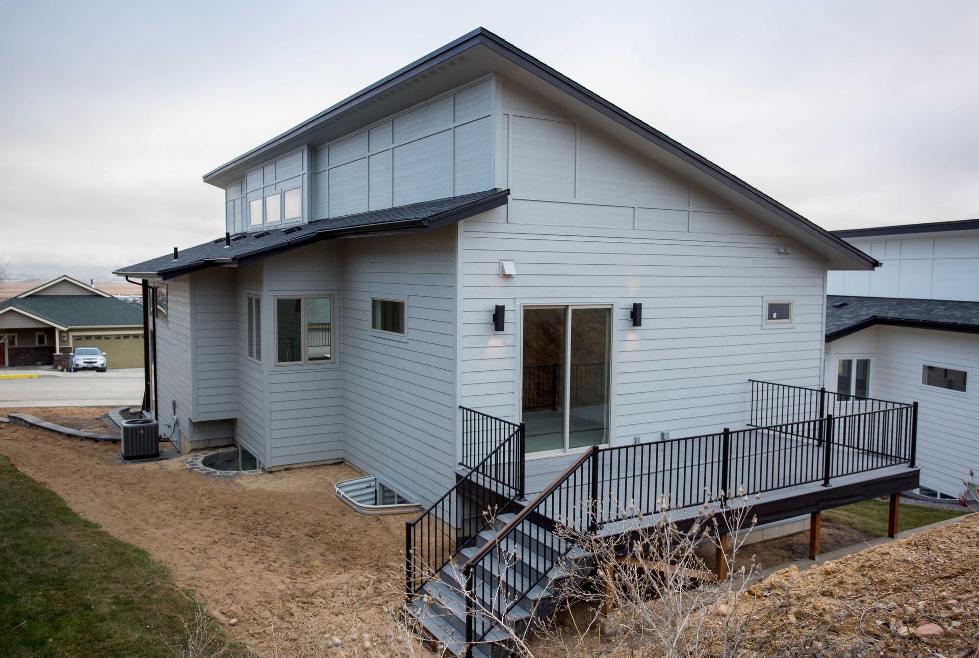 A modern two-story light gray house with a deck, stairs, and a black roof in a residential neighborhood.