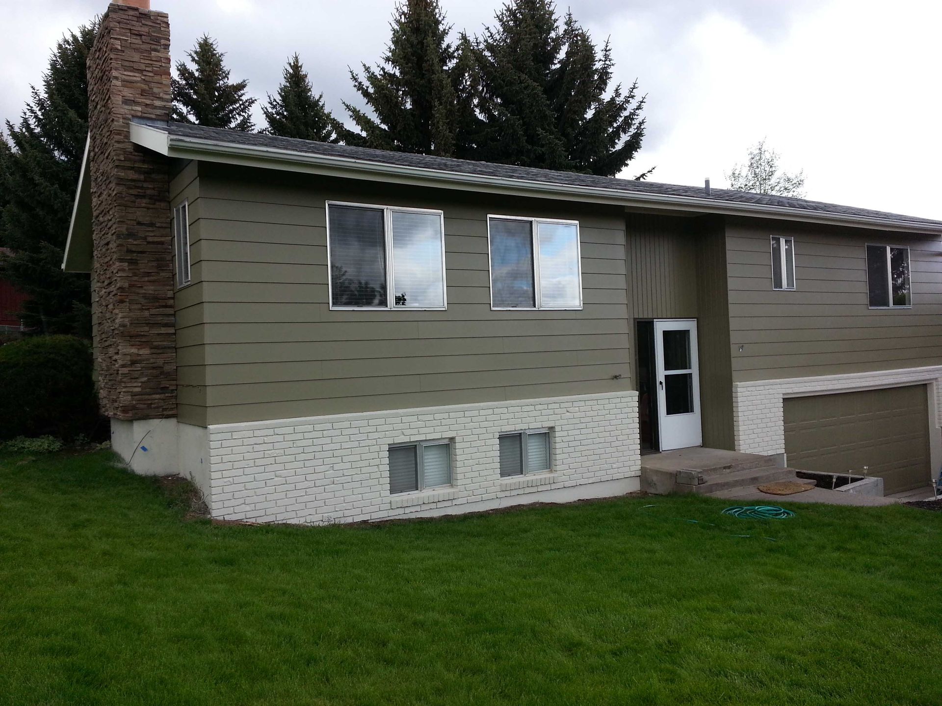A split-level home with olive-green siding and a white brick lower exterior, stone chimney, and an attached garage.