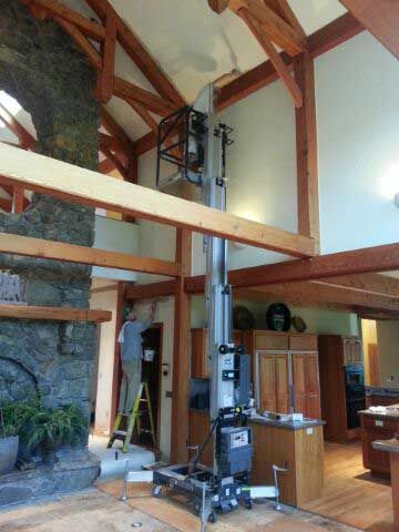 A person uses a vertical lift to reach the high ceiling of a room with exposed timber beams and a stone fireplace.