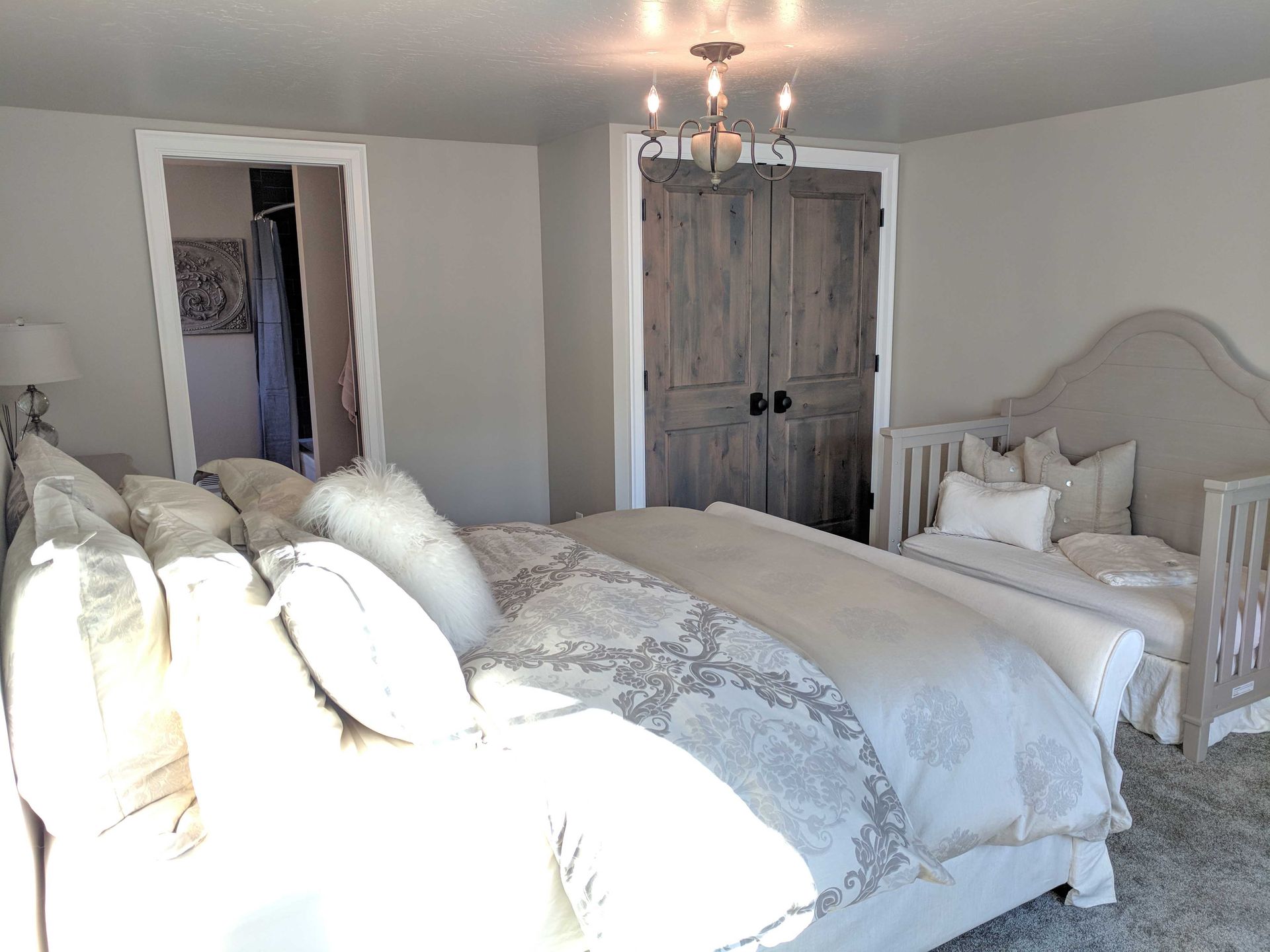 A bedroom features a large bed with neutral bedding, a small upholstered daybed, a wooden closet, and a light fixture.
