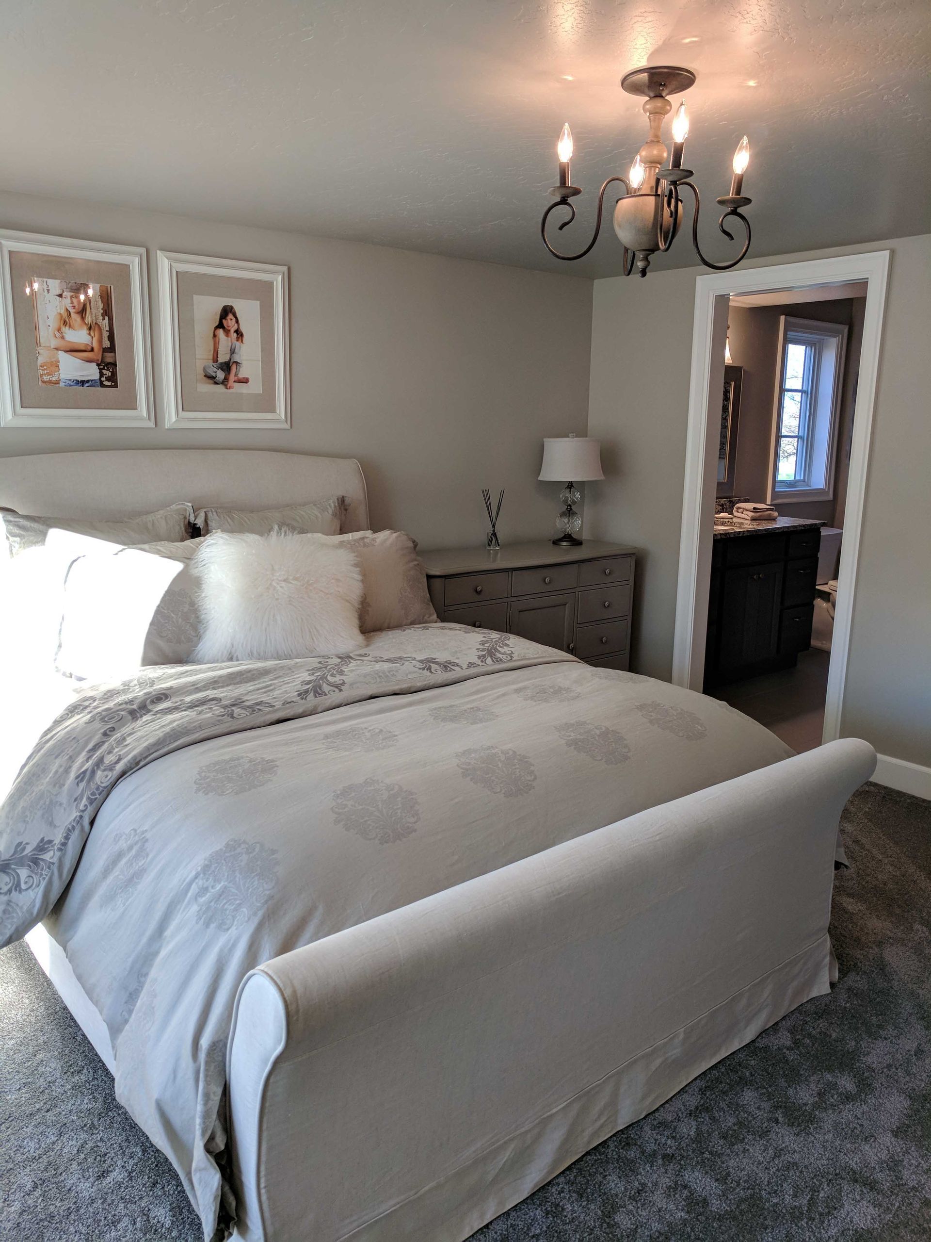 A neutrally decorated bedroom featuring a beige upholstered bed, grey bedding, two framed wall photos, and a chandelier.