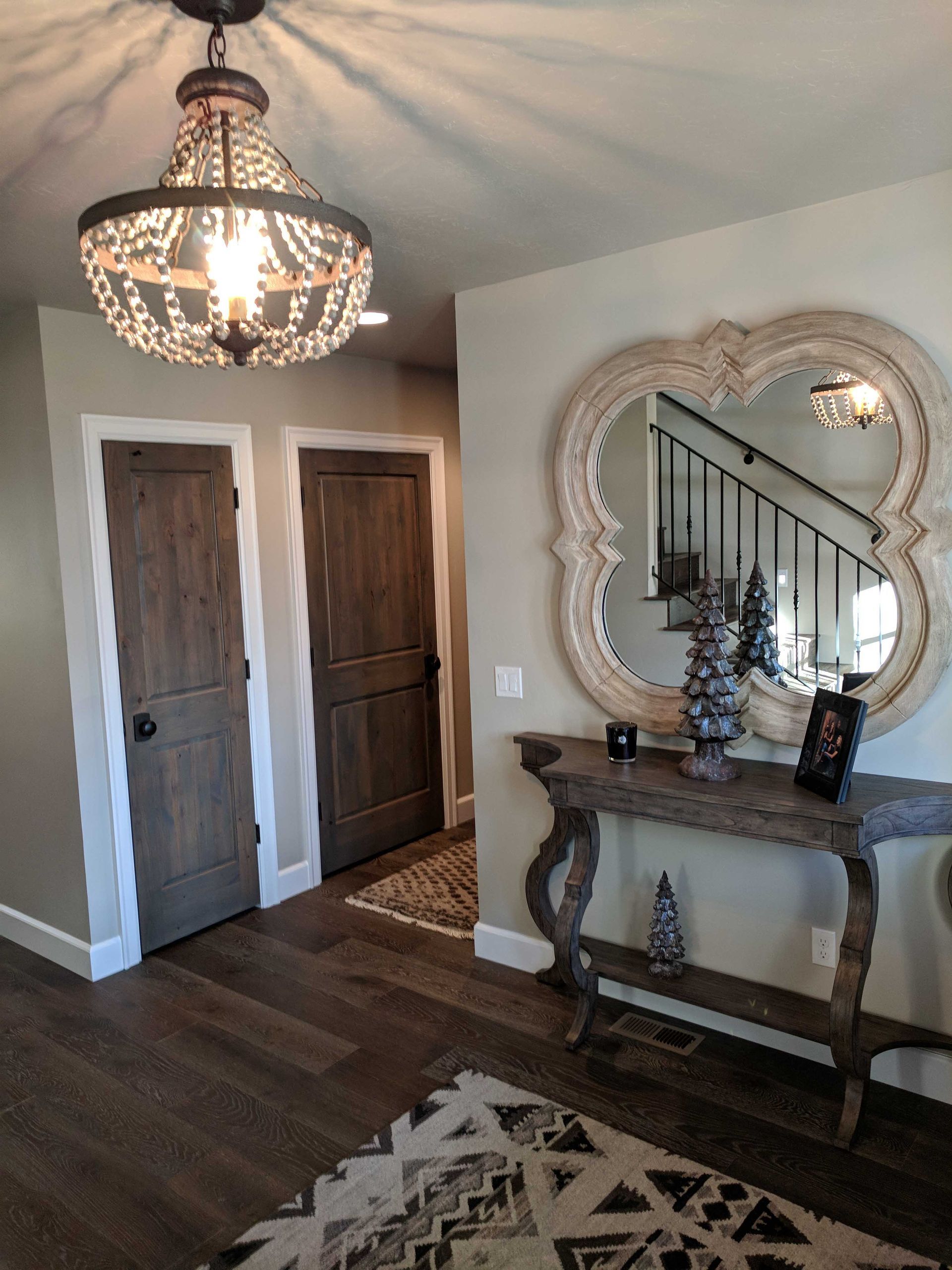 A wooden console table under a decorative mirror in a hallway with dark wood doors, patterned rug, and bead chandelier.