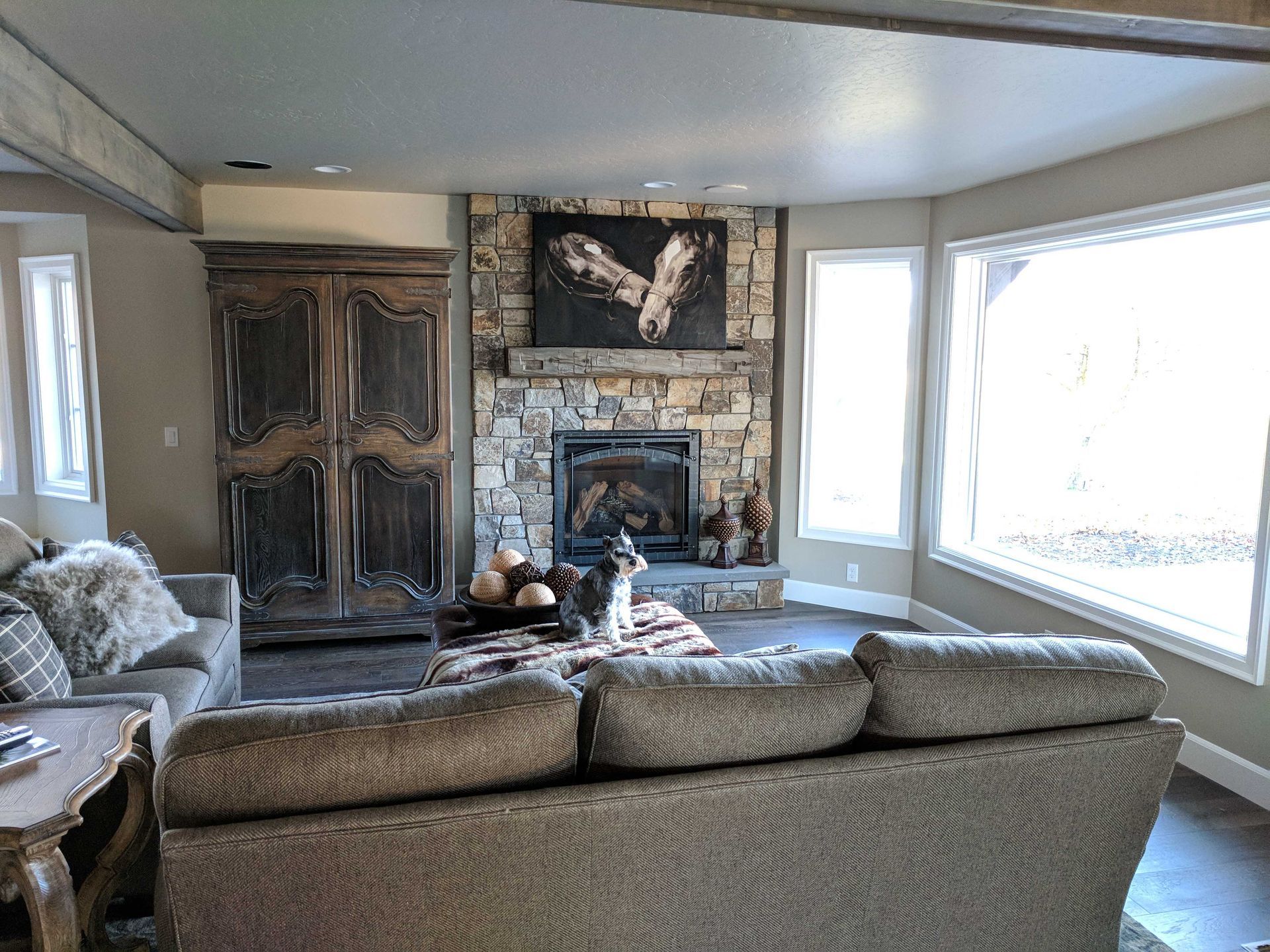 A living room with a stone fireplace, a large wooden armoire, a gray sofa, and a small dog standing on a patterned ottoman.