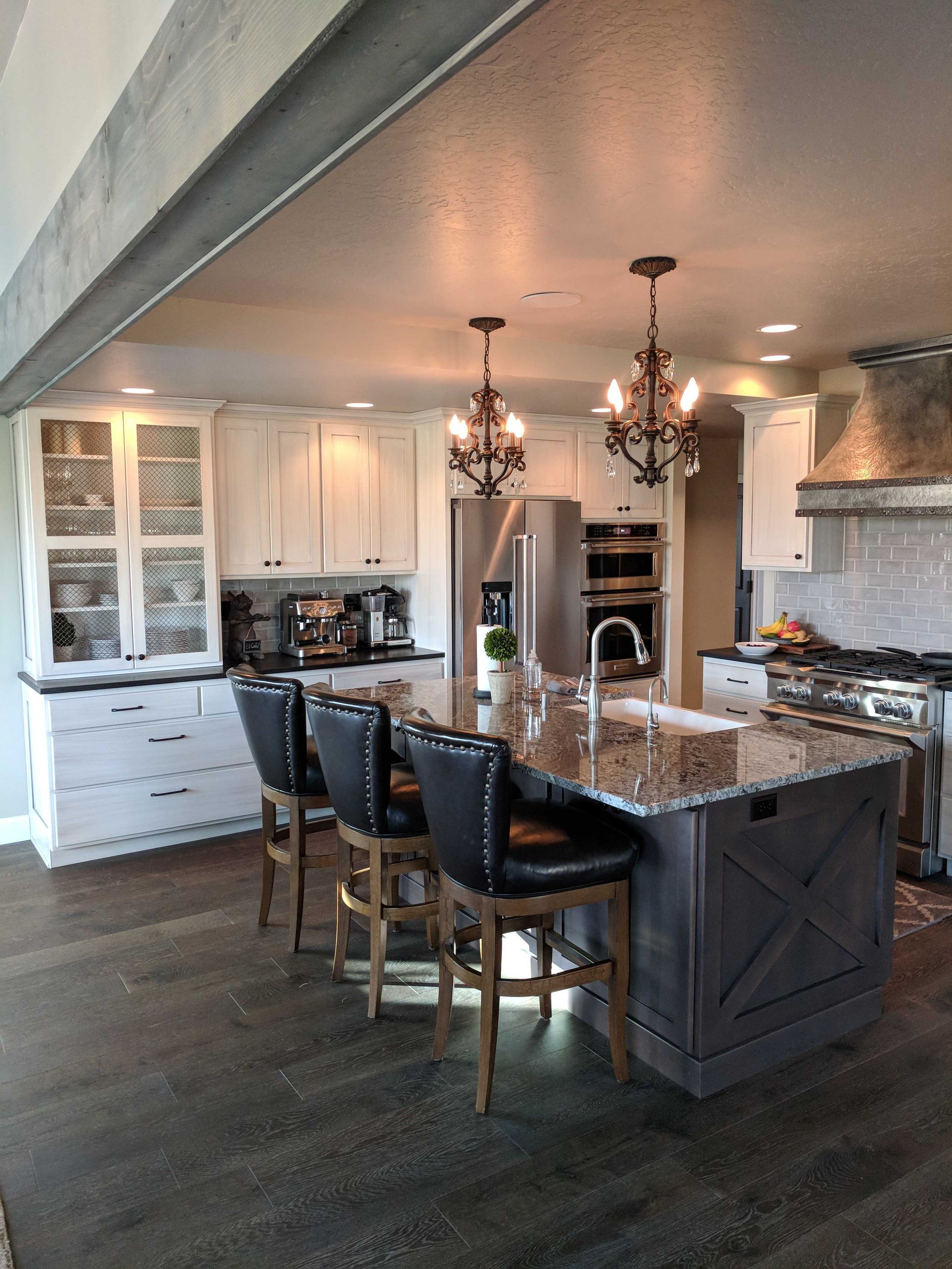 A bright kitchen with white cabinetry, a dark gray island with three black stools, and two ornate hanging light fixtures.