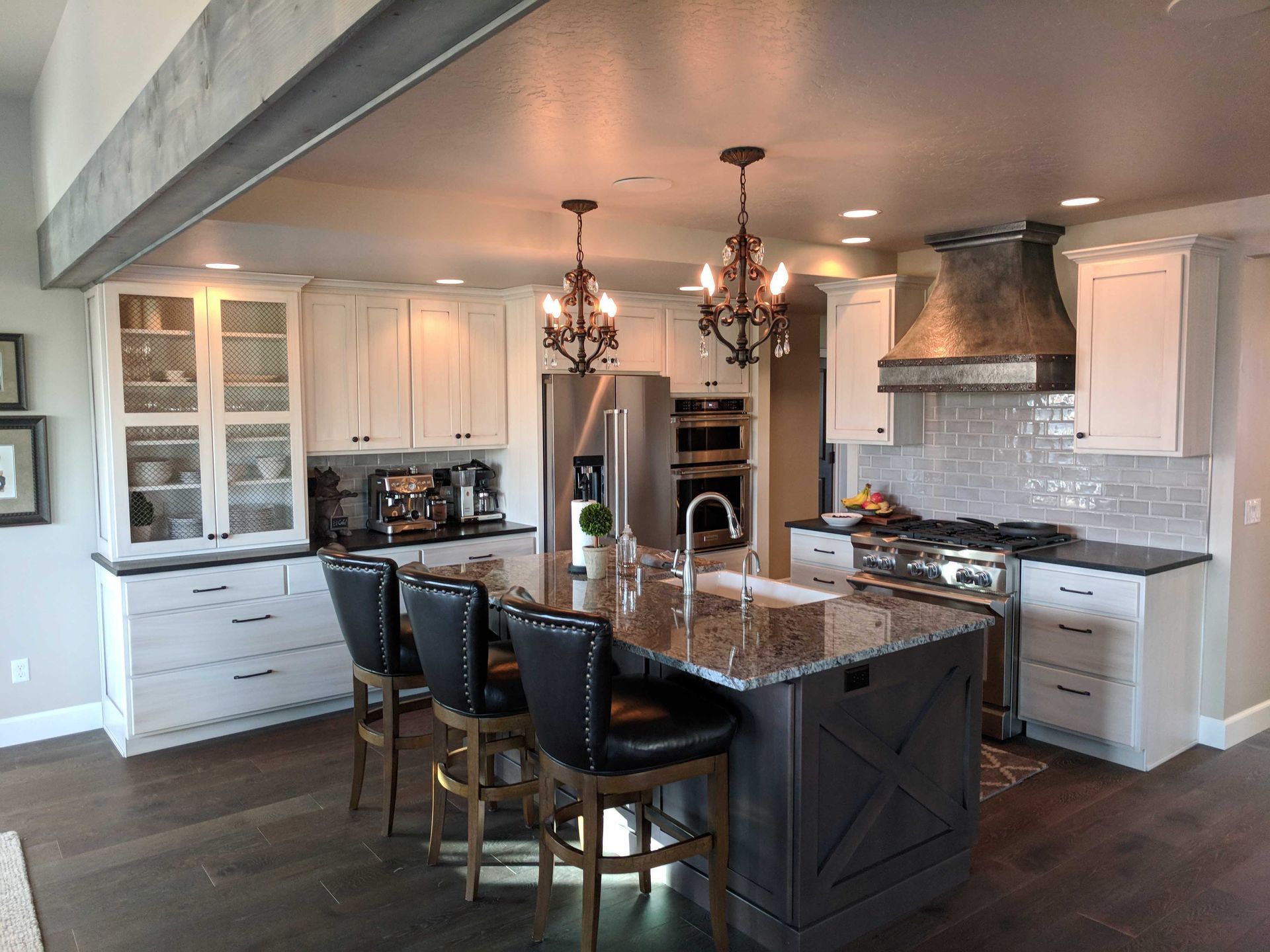 A kitchen features a granite-topped island with three black stools, white cabinetry, a metal range hood, and wood floors.