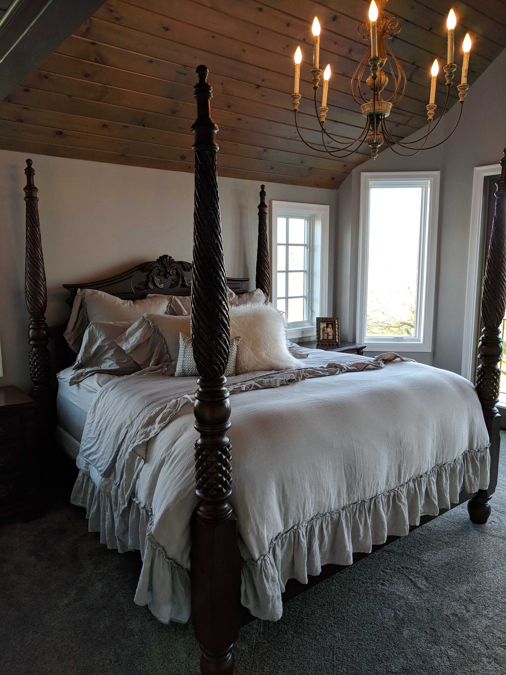 A four-poster wooden bed with light gray bedding sits in a bedroom with a sloped wood ceiling and a candle chandelier.