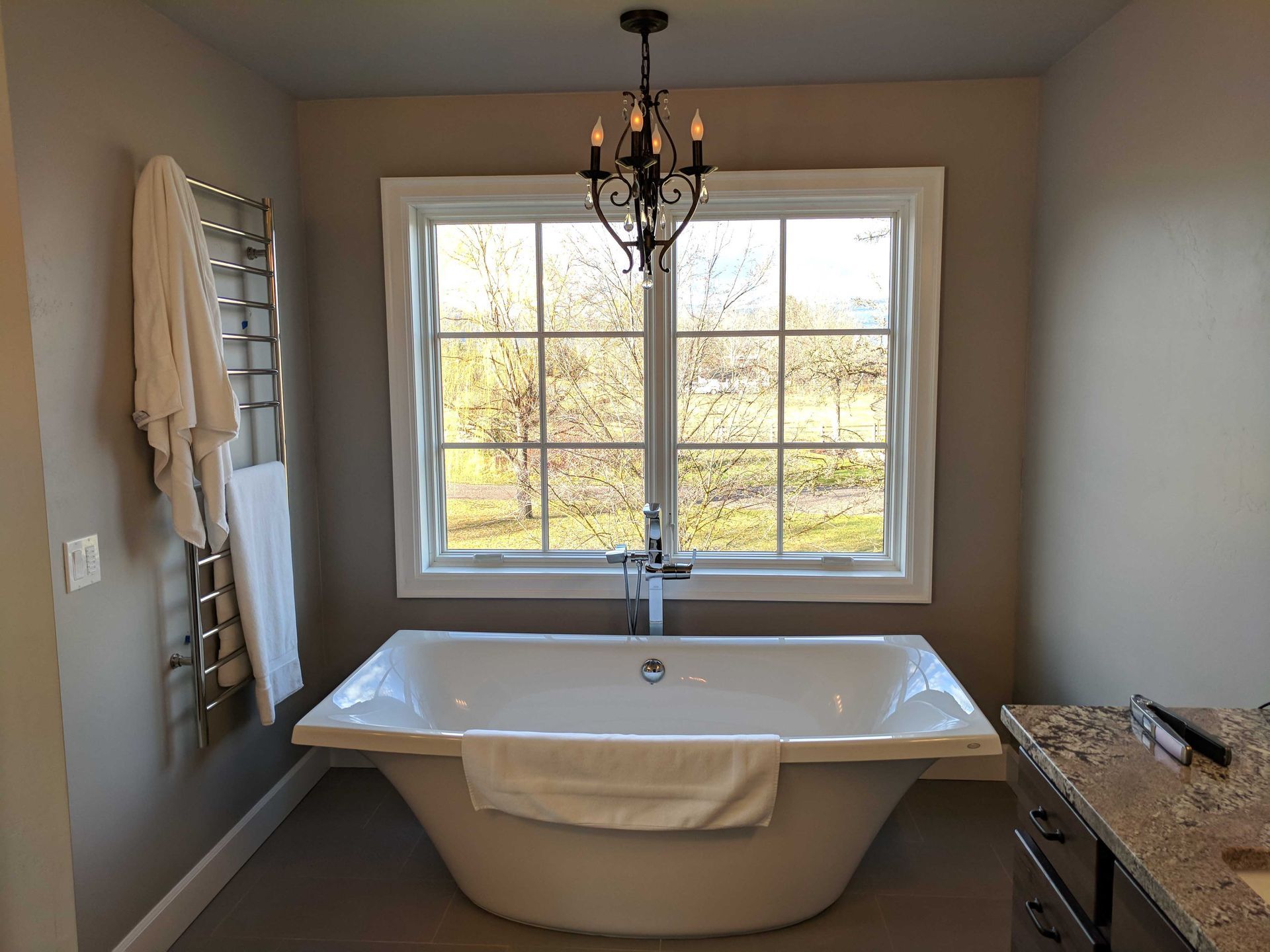 A white freestanding bathtub sits beneath a window in a bathroom with grey walls and a dark chandelier.