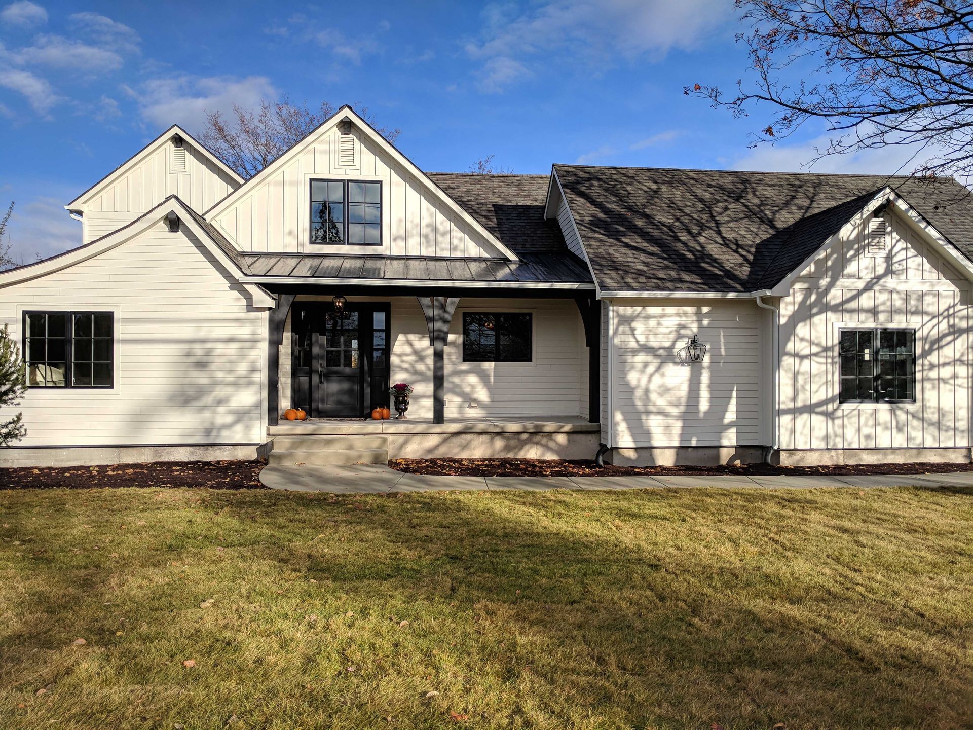 A white farmhouse with a metal porch roof, dark window frames, and a brown shingled roof under a bright blue sky.