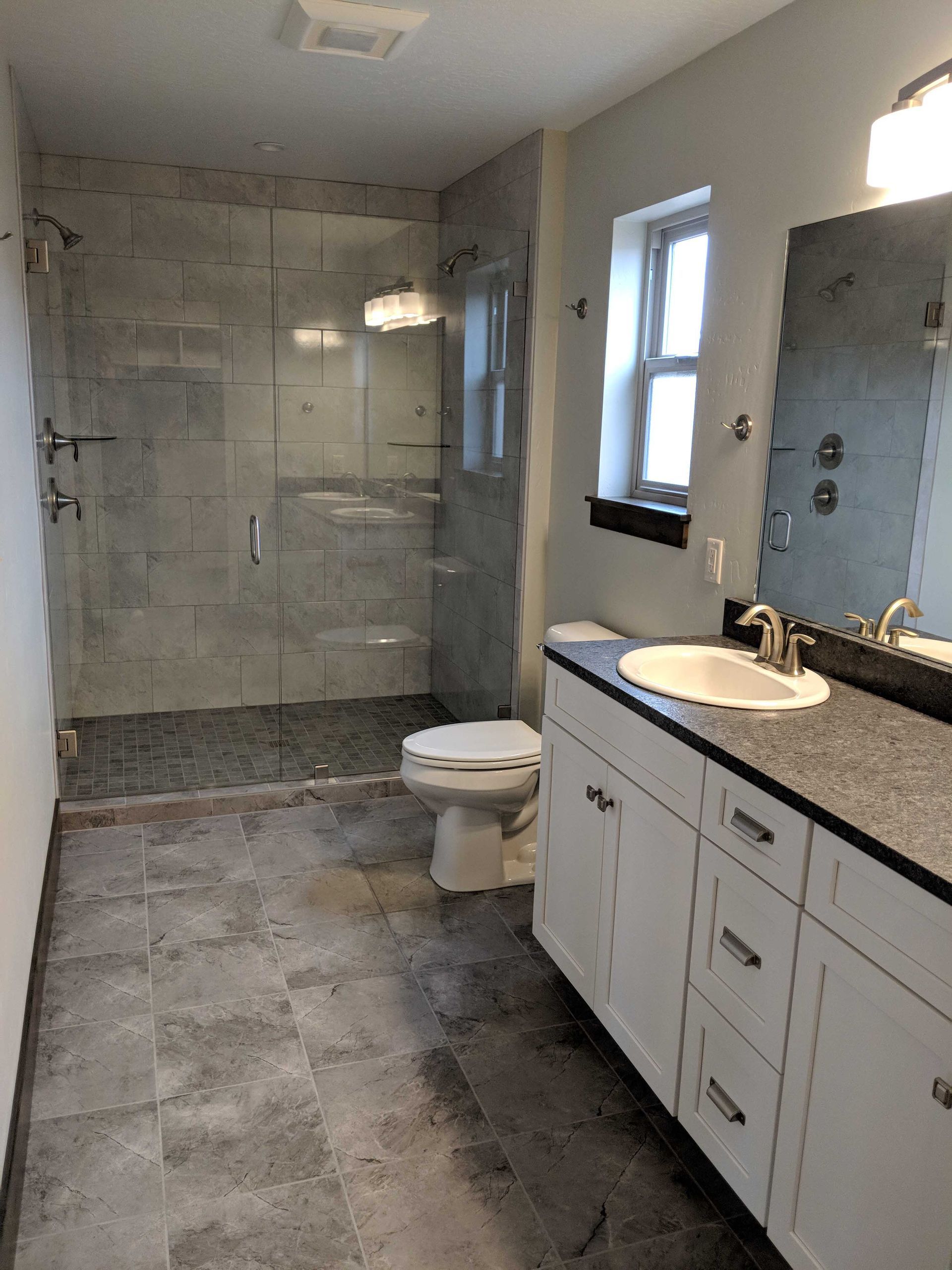 A modern bathroom features a glass-enclosed shower, a white vanity with a dark countertop, and grey tile flooring.