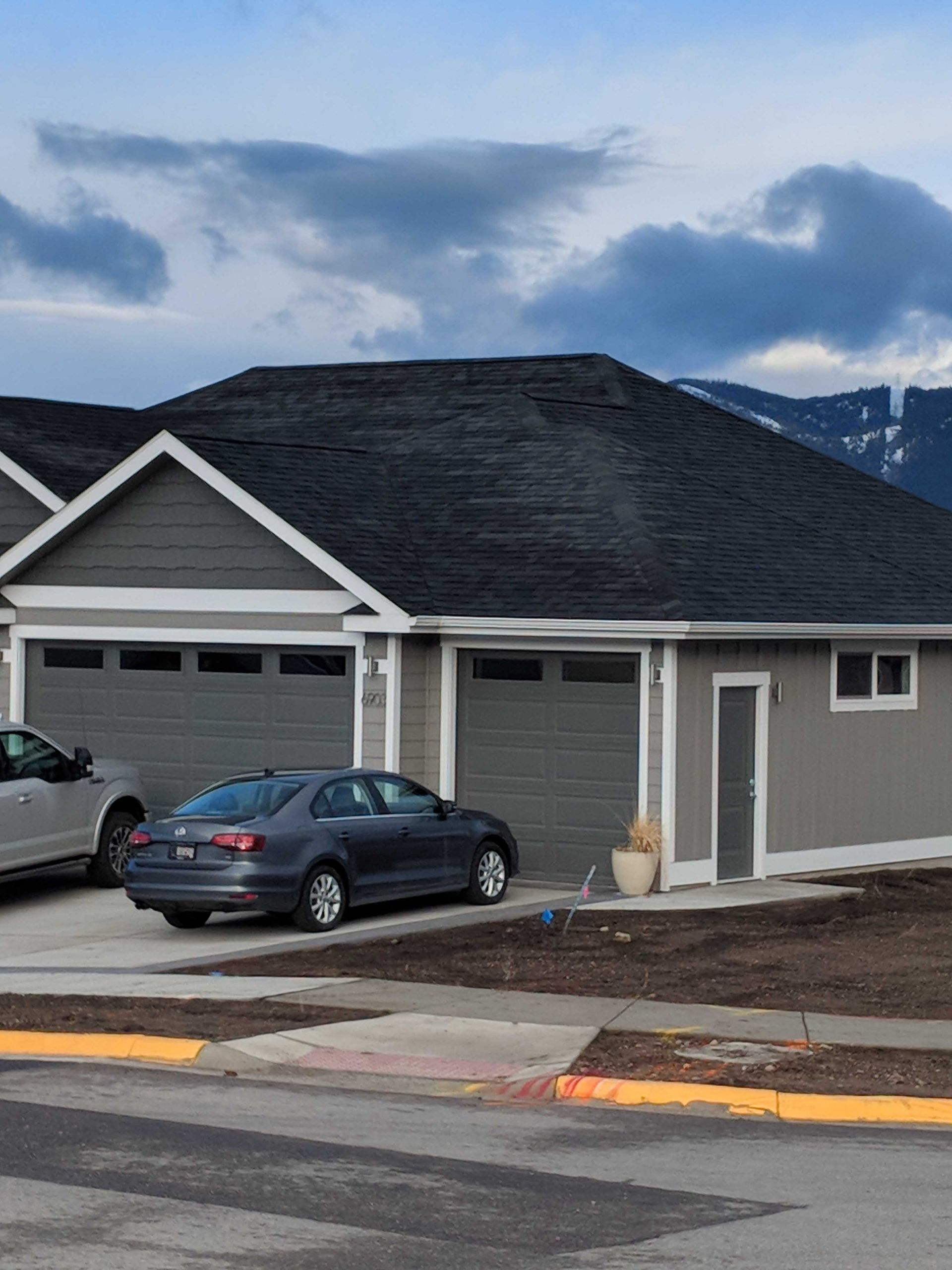 A gray house with two garage doors and a car parked in front, set against a backdrop of clouds and distant mountains.