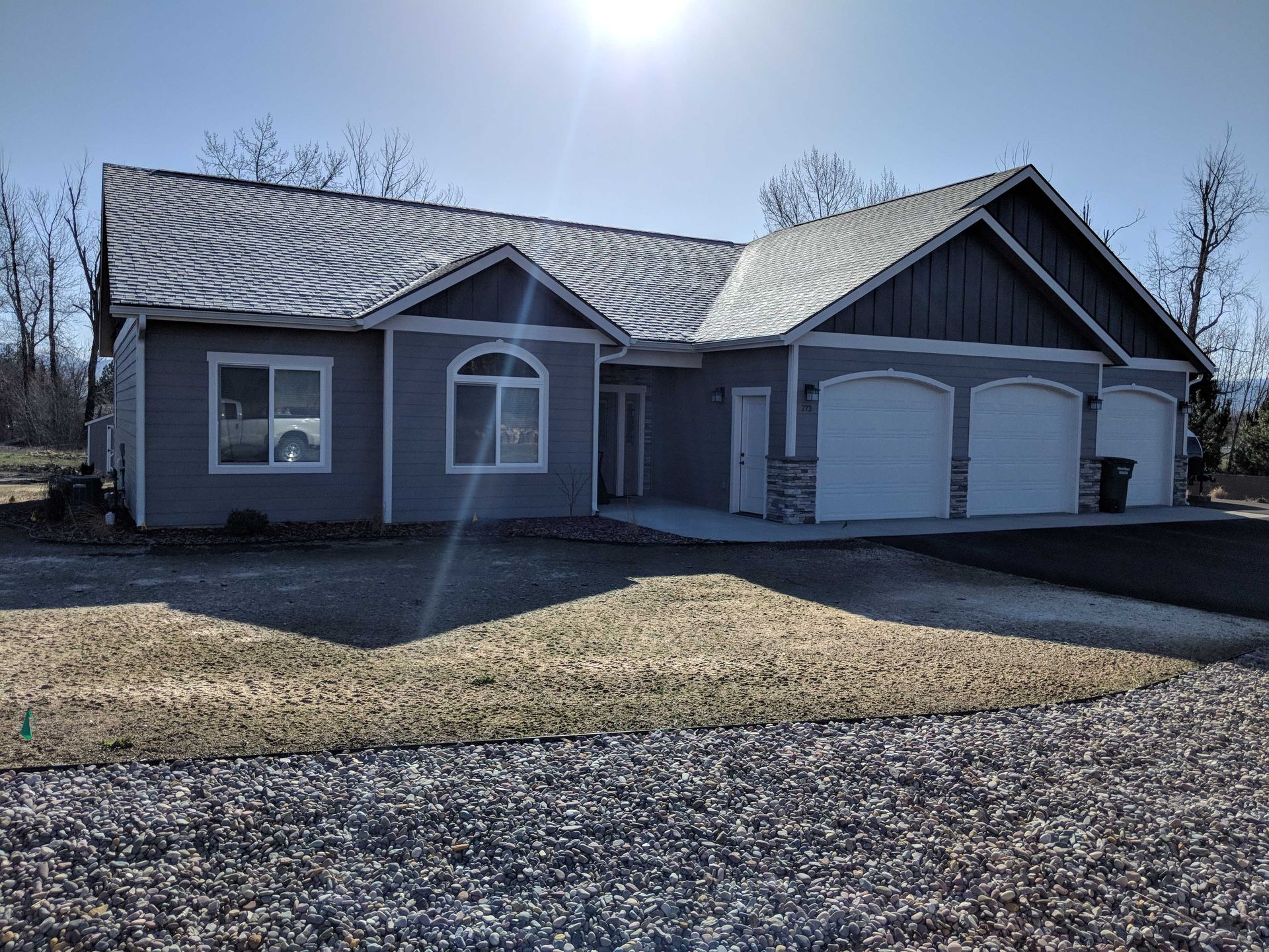 A grey, single-story house with a three-car garage, stone accents, and a large gravel driveway under a bright blue sky.