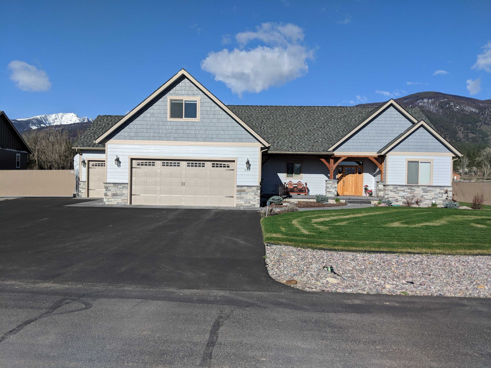 A light blue, single-story house with a garage and a stone foundation against a mountain backdrop under a blue sky.