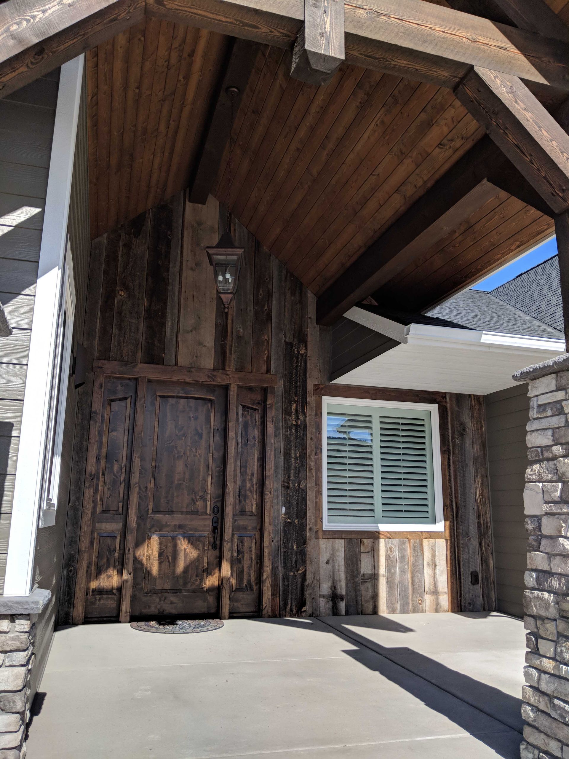 Front entrance of a home with a large dark wood door, stone pillars, and wood-paneled porch ceiling.