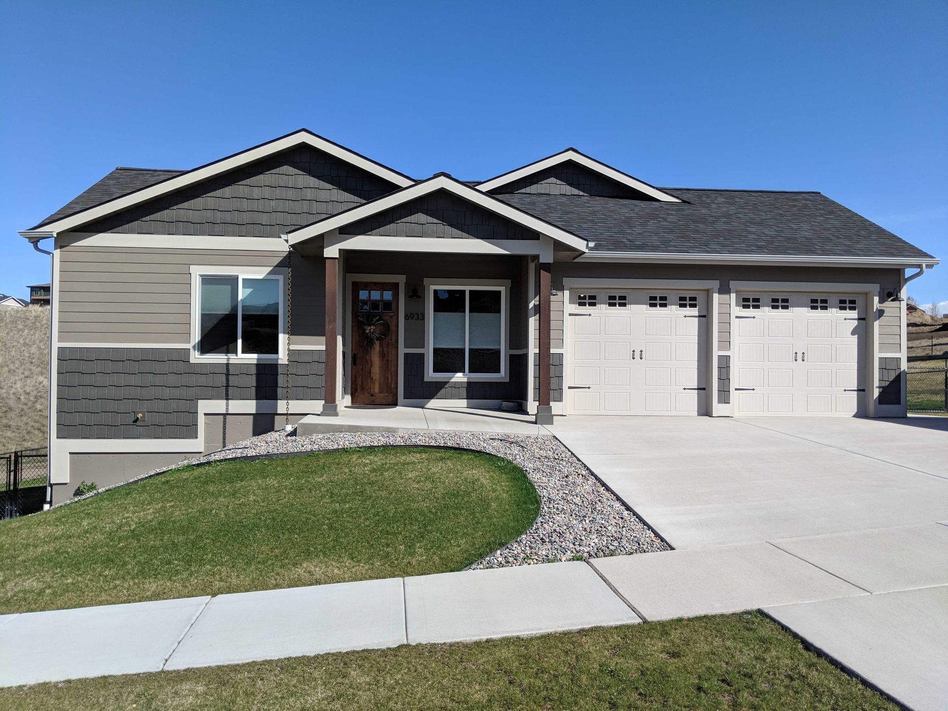 A beige and grey ranch-style house with a two-car garage, front porch, and a curved lawn under a clear blue sky.