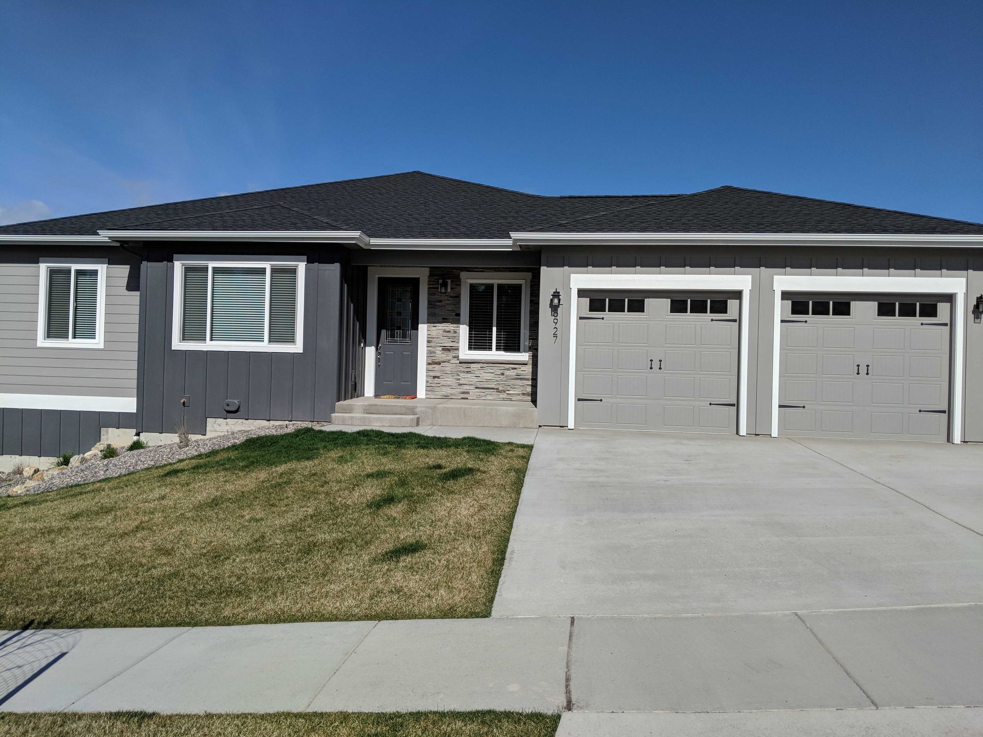 A modern single-story suburban house with a gray exterior, stone accents, two garage doors, and a front lawn.