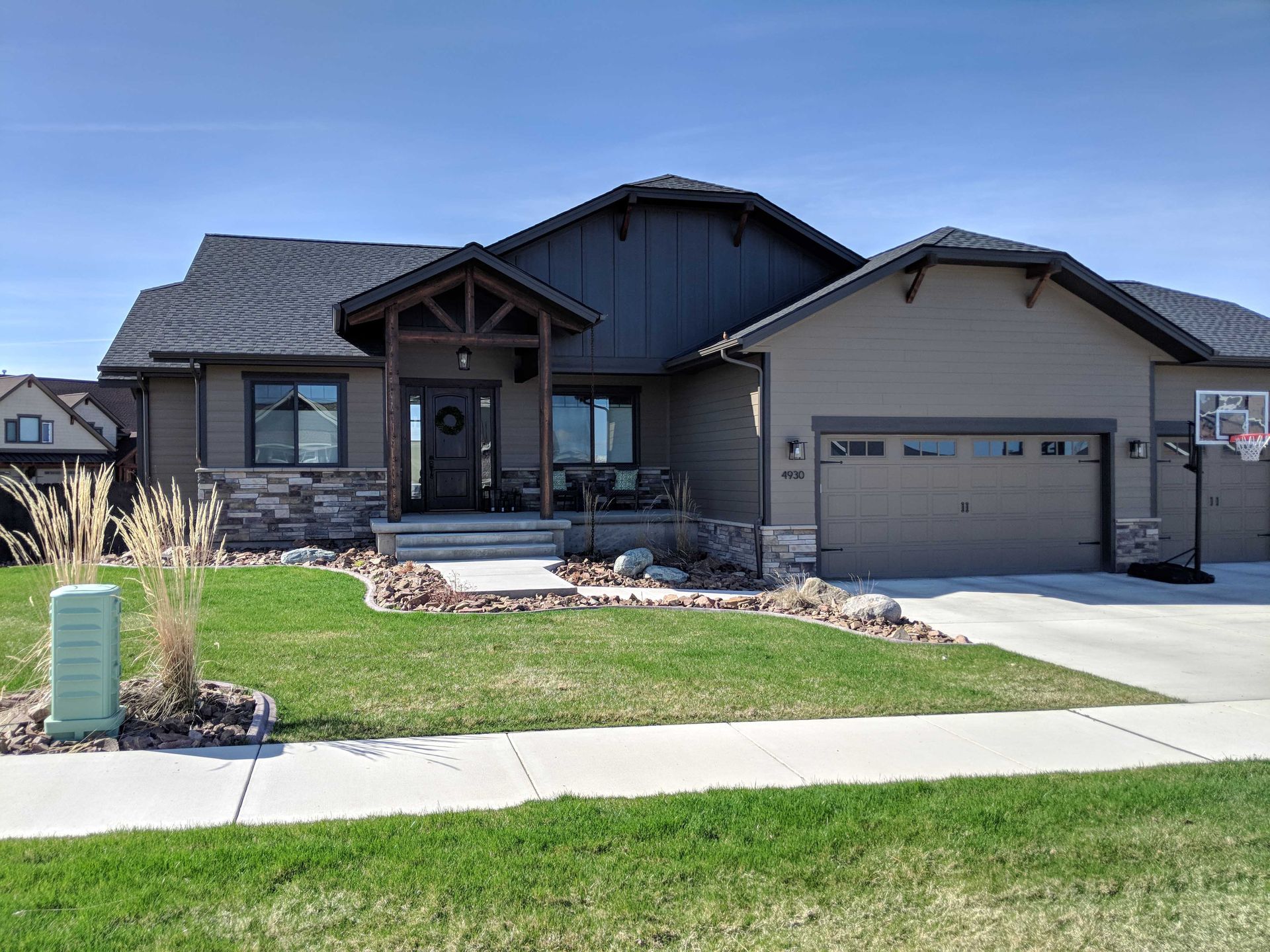Single-story suburban house with a dark gabled roof, tan siding, stone accents, a wood-trimmed porch, and a garage.