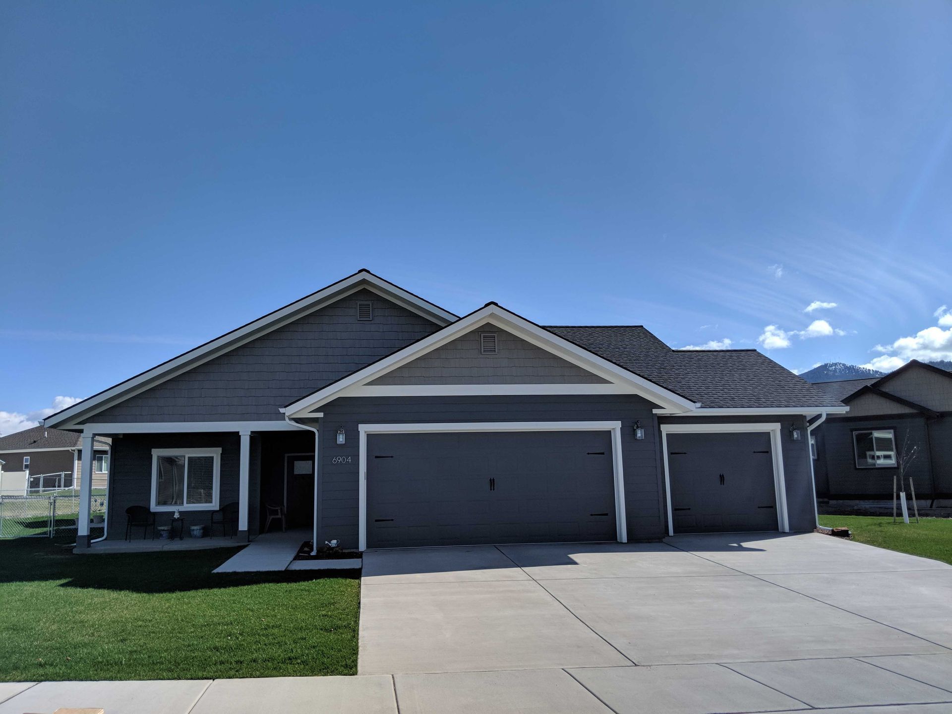 A gray, single-story suburban house with a two-car garage, a covered front porch, and a grassy front lawn under a blue sky.