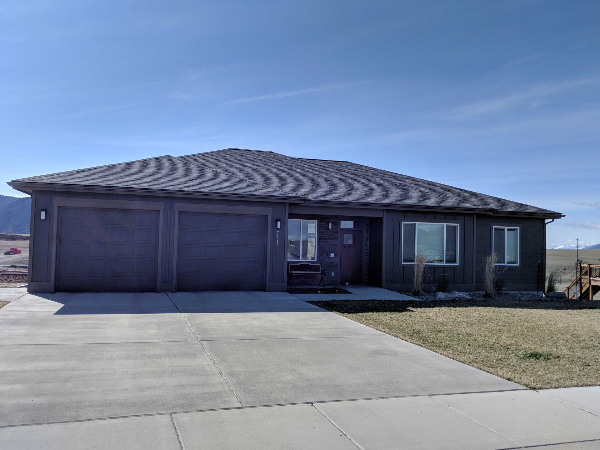 A single-story, dark gray suburban house with a two-car garage and a concrete driveway under a clear blue sky.