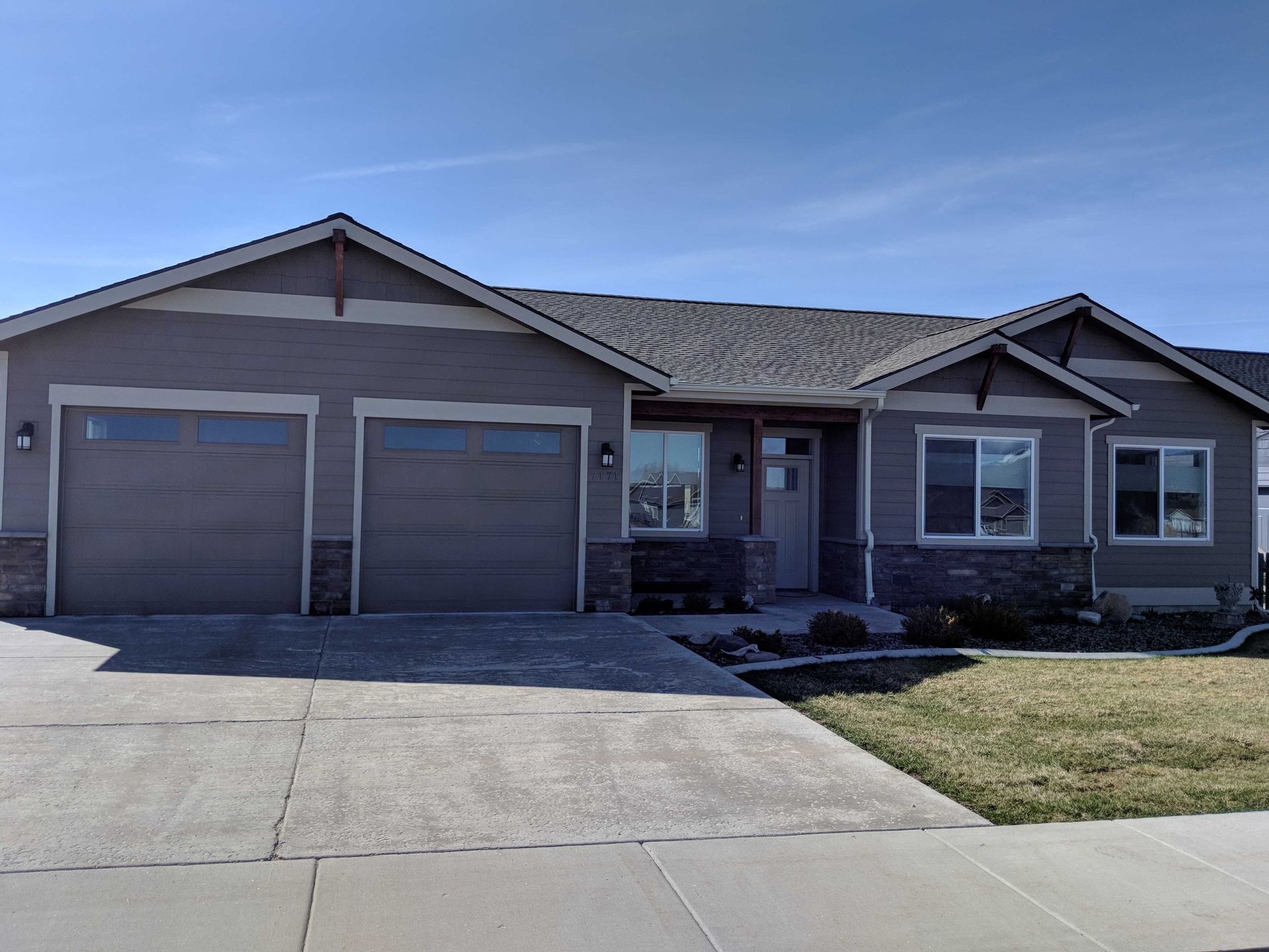 Single-story house with a two-car garage, gray siding, stone accents, and a front porch under a clear blue sky.
