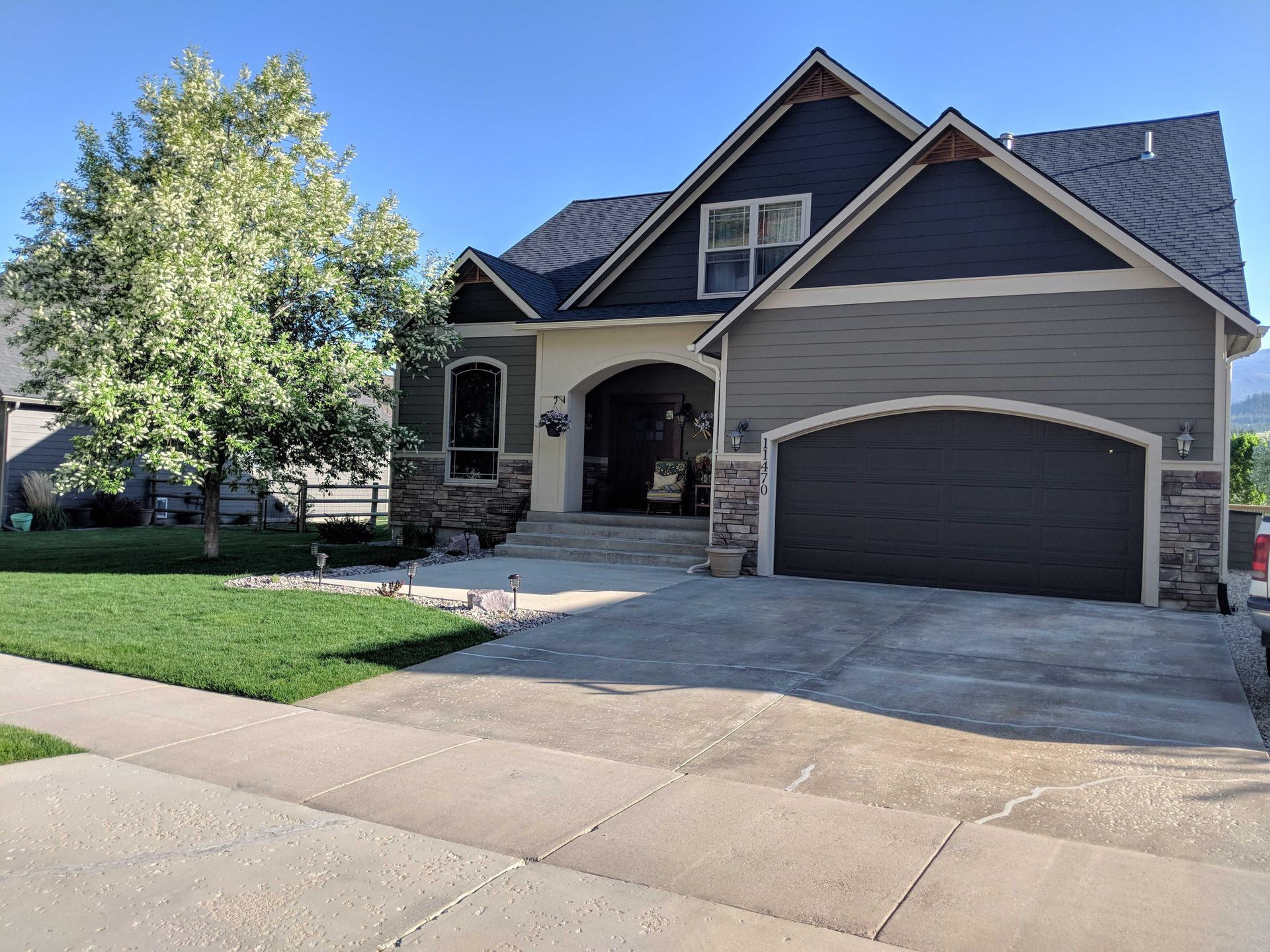 A two-story suburban home with a dark gray garage, blue siding, stone accents, and a large front tree under a blue sky.