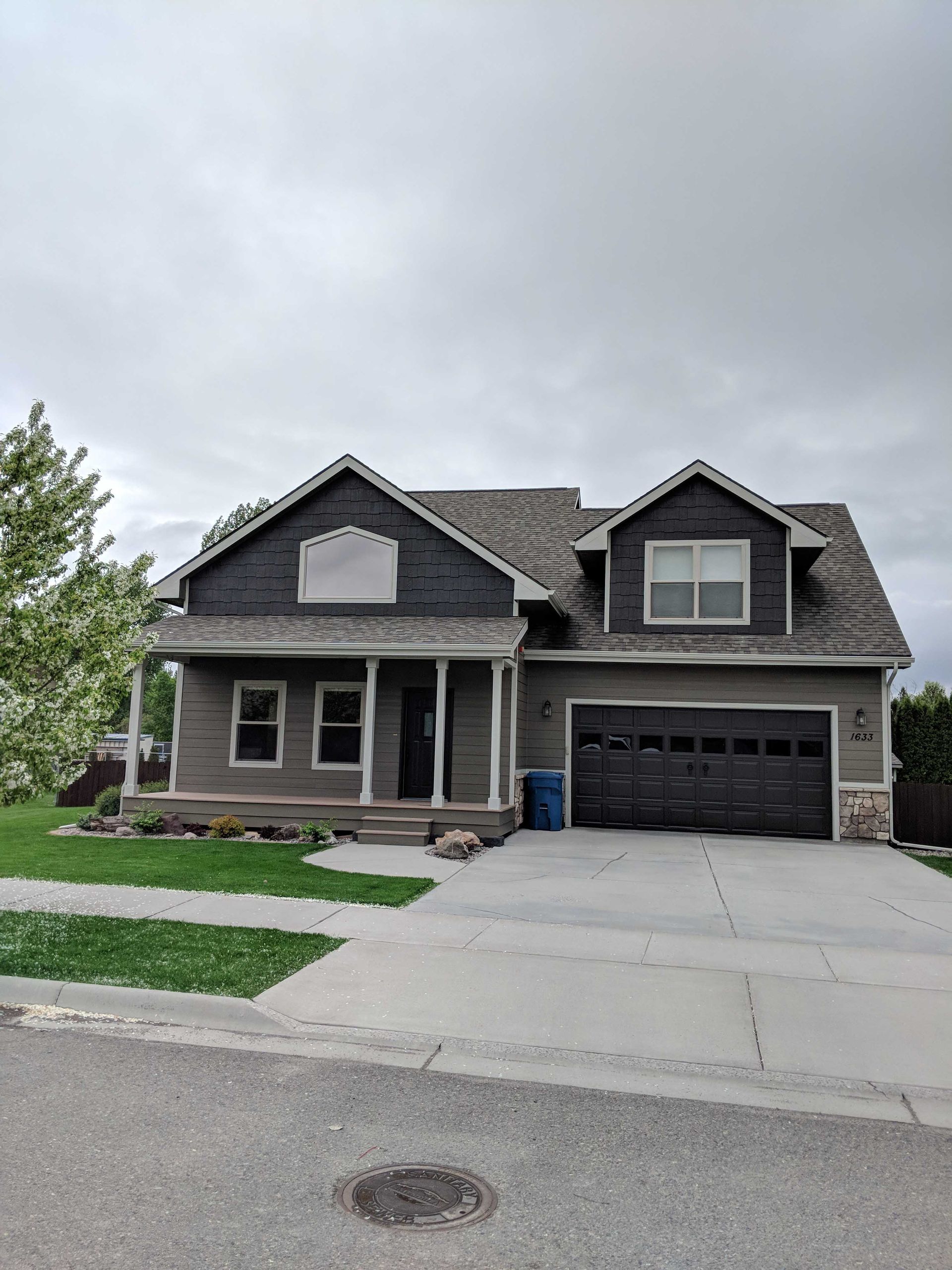 A two-story grey house with dark trim, a covered front porch, a two-car garage, and a green front lawn under a cloudy sky.