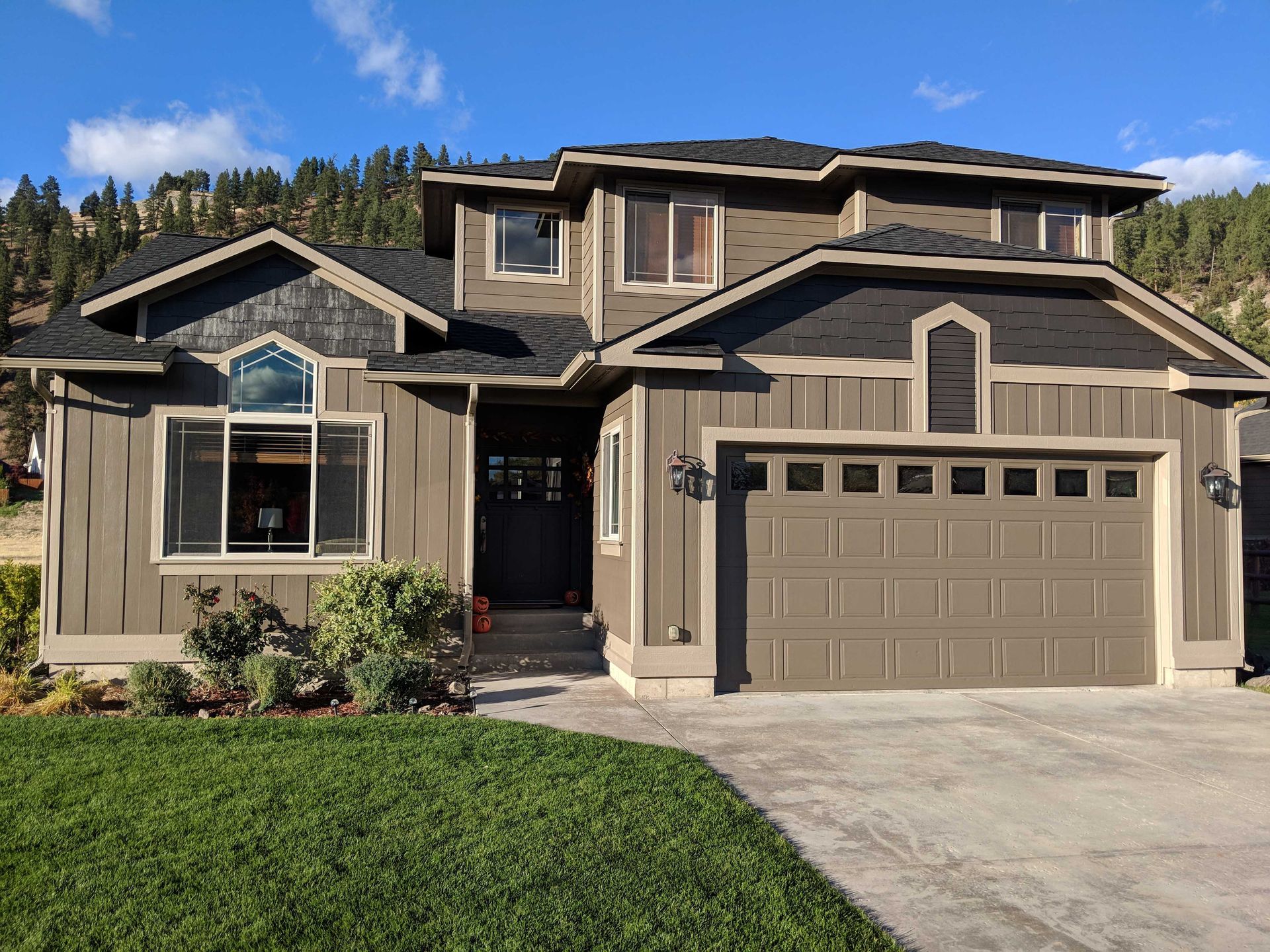 A two-story taupe house with dark brown trim, a large garage, and an arched window, set against a tree-lined hillside.