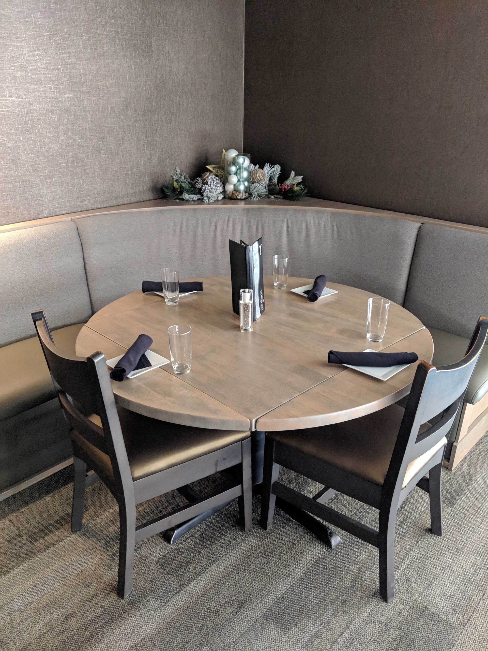 A restaurant booth featuring a round wooden table, two chairs, and a curved cushioned seat in a corner with neutral tones.
