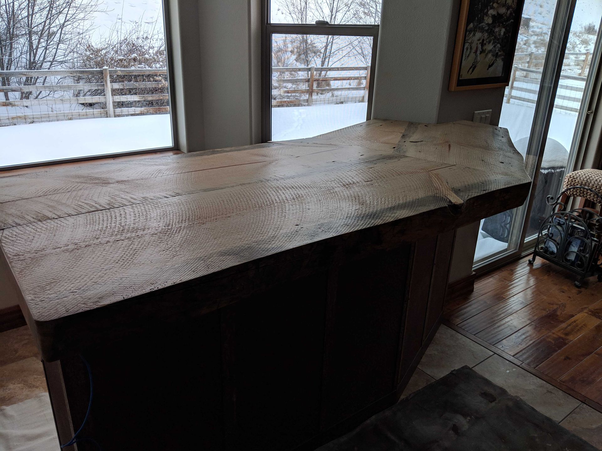 A wooden kitchen island with a natural, unfinished edge in a bright room with windows overlooking a snowy landscape.