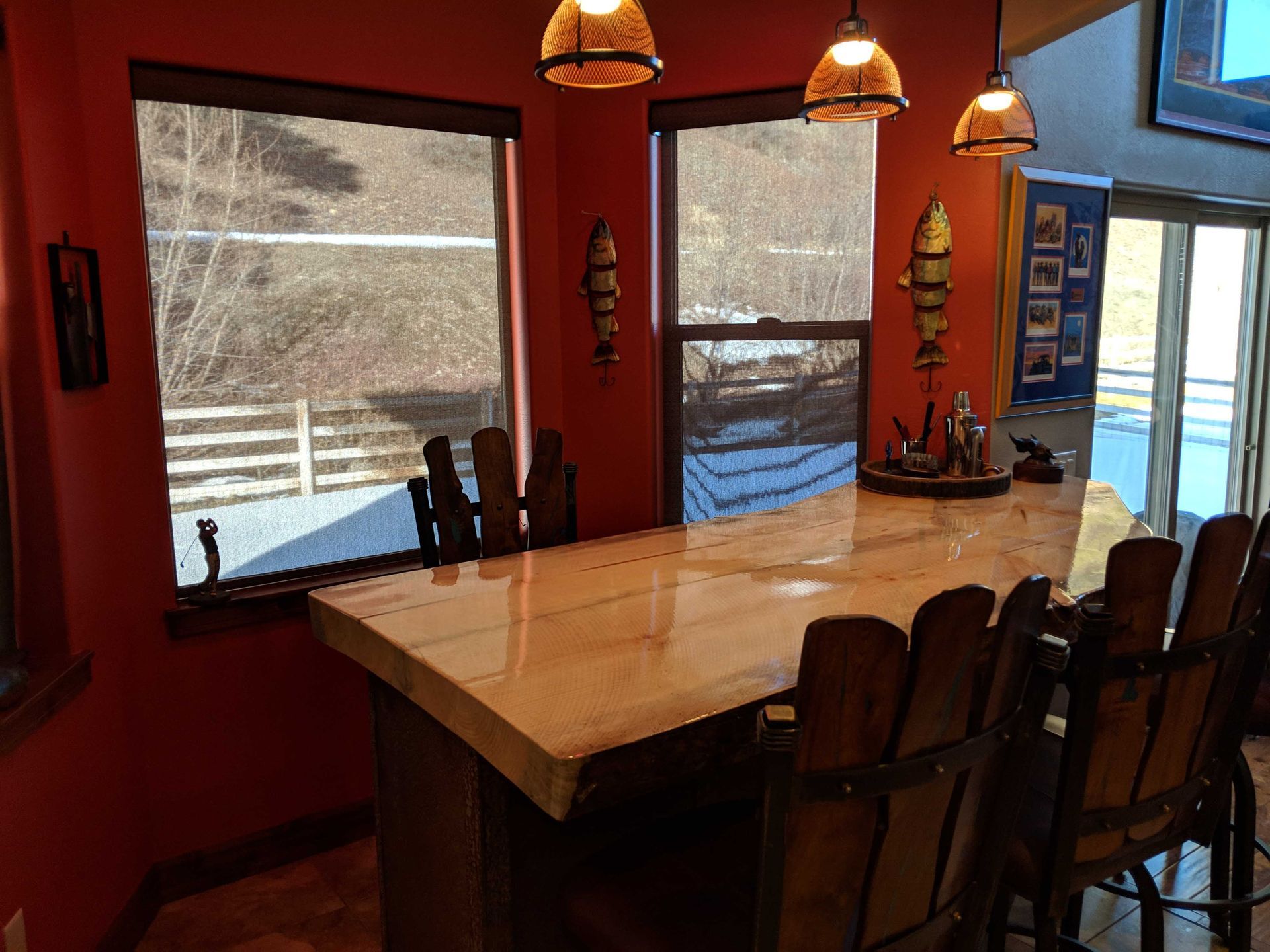 A wooden bar counter with tall rustic chairs sits in a room with vibrant red walls, windows, and hanging pendant lights.