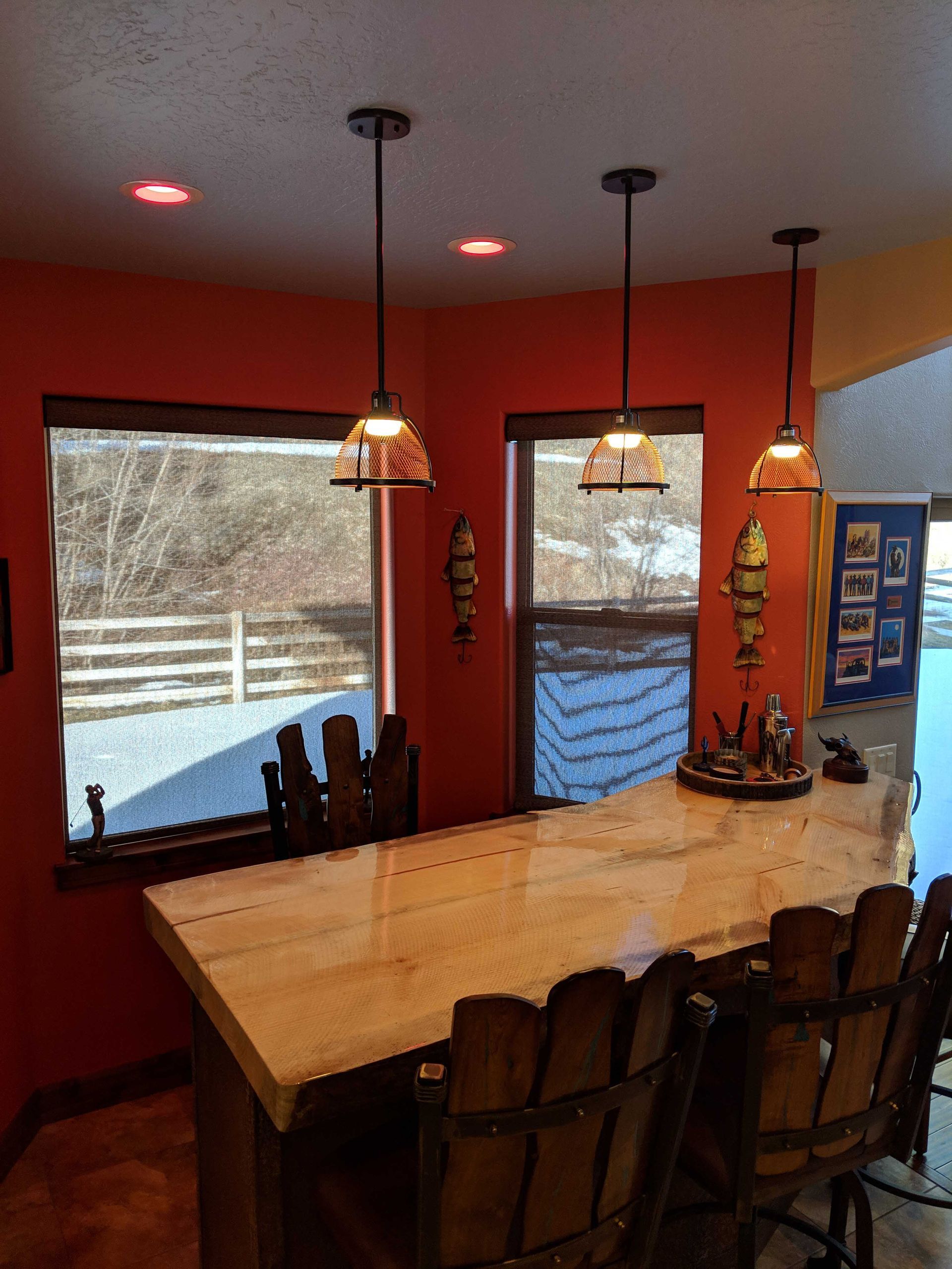 A high wooden bar counter with three hanging pendant lights in front of large windows and rust-colored walls.