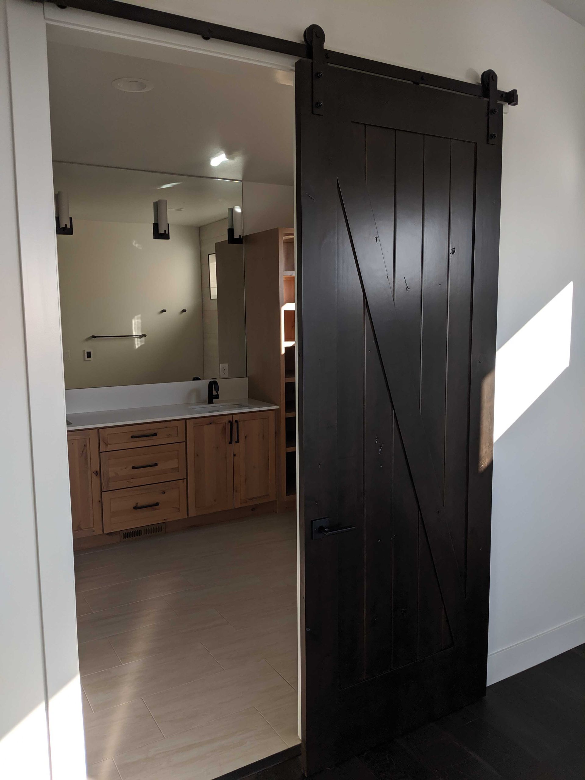 A dark-stained wooden barn door with diagonal bracing slides open to reveal a bathroom with a wooden vanity and mirror.