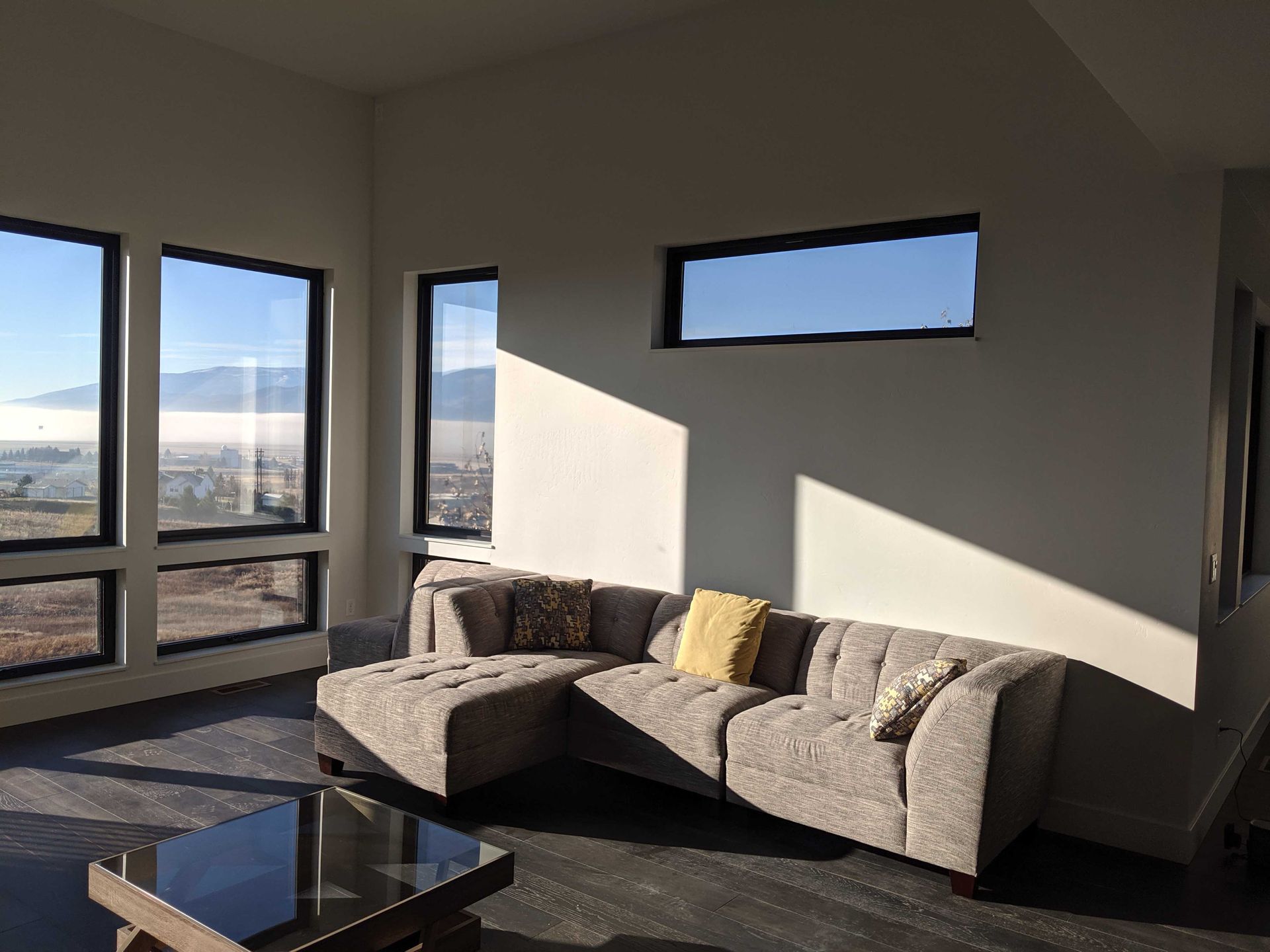 A modern living room with a gray sectional sofa, a coffee table, and large windows revealing a scenic, misty landscape.