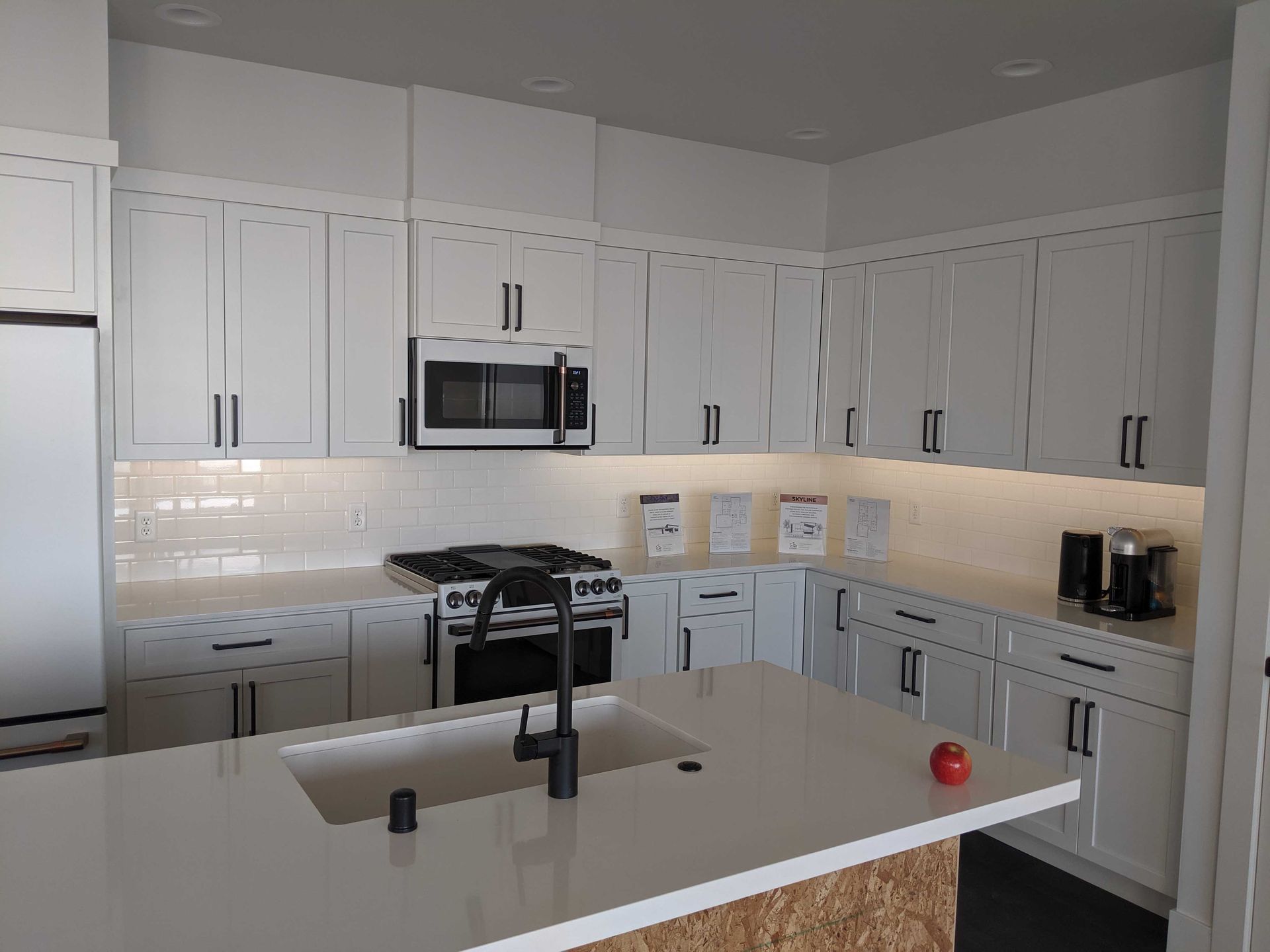 A modern white kitchen featuring an island with a sink, white cabinets, a microwave over a gas stove, and an apple.