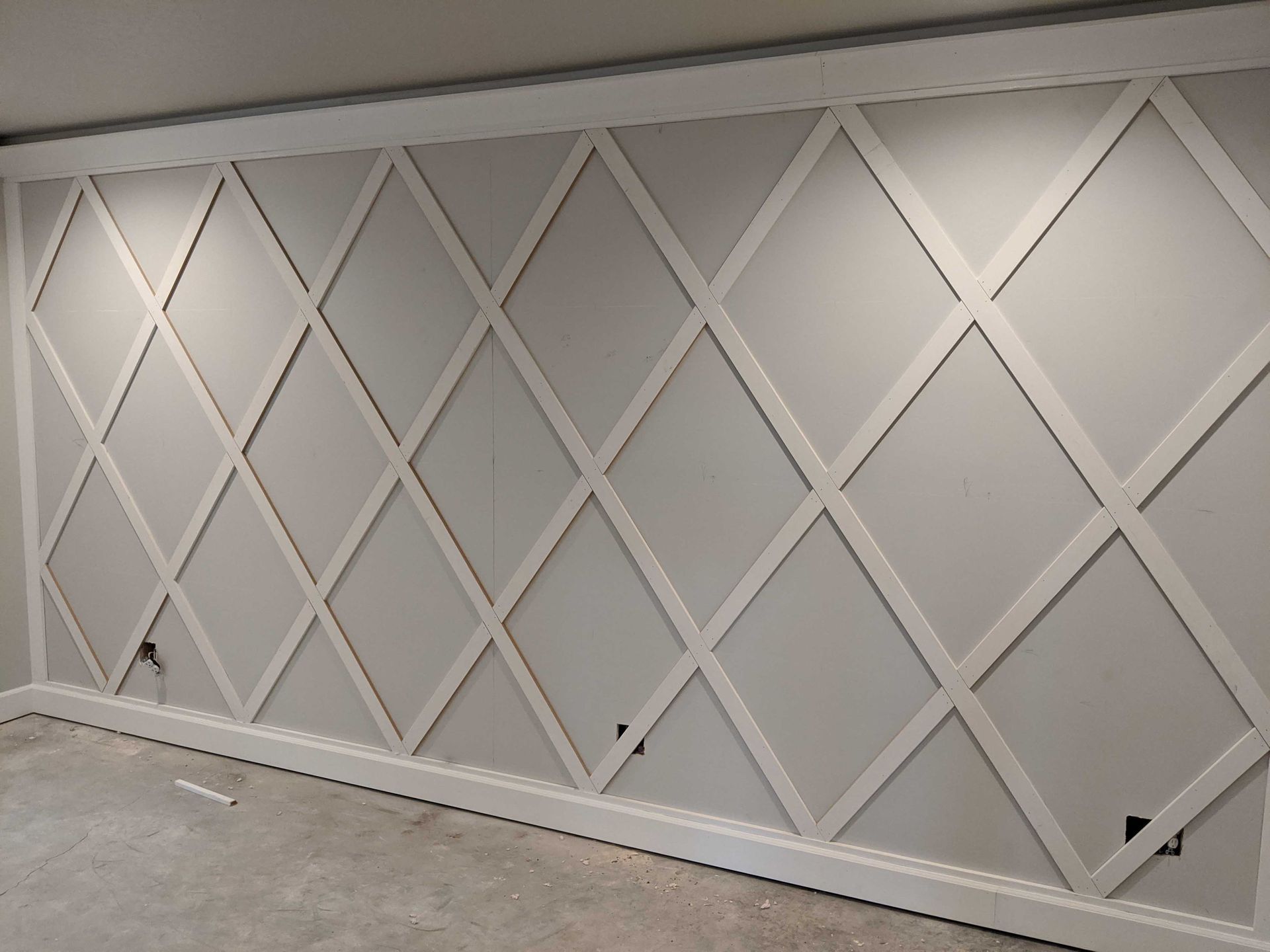 A wall feature with white wooden lattice molding creating a diamond pattern over a light gray wall surface.