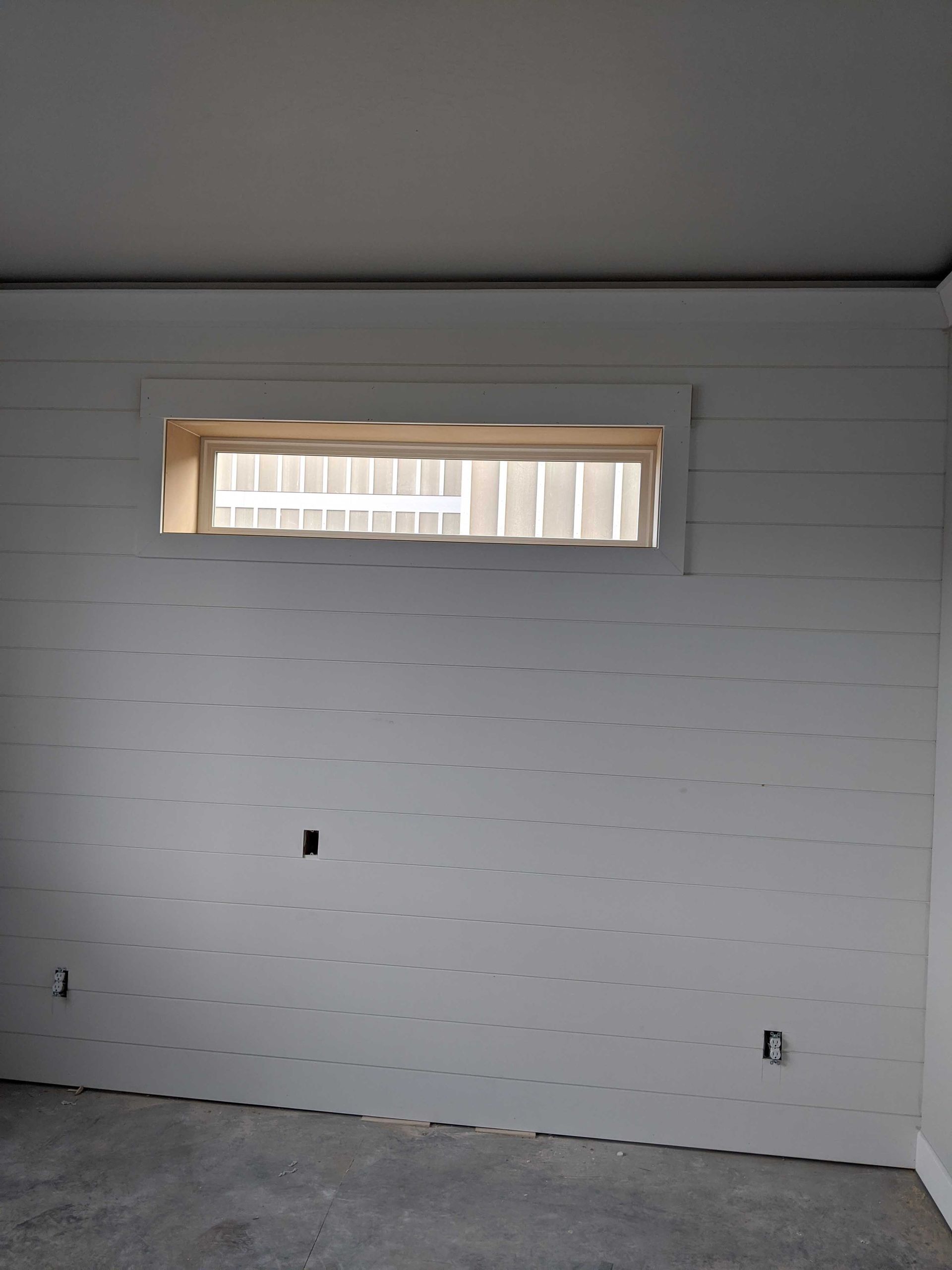 A white shiplap wall featuring a rectangular horizontal window near the ceiling, with visible electrical wall outlets.