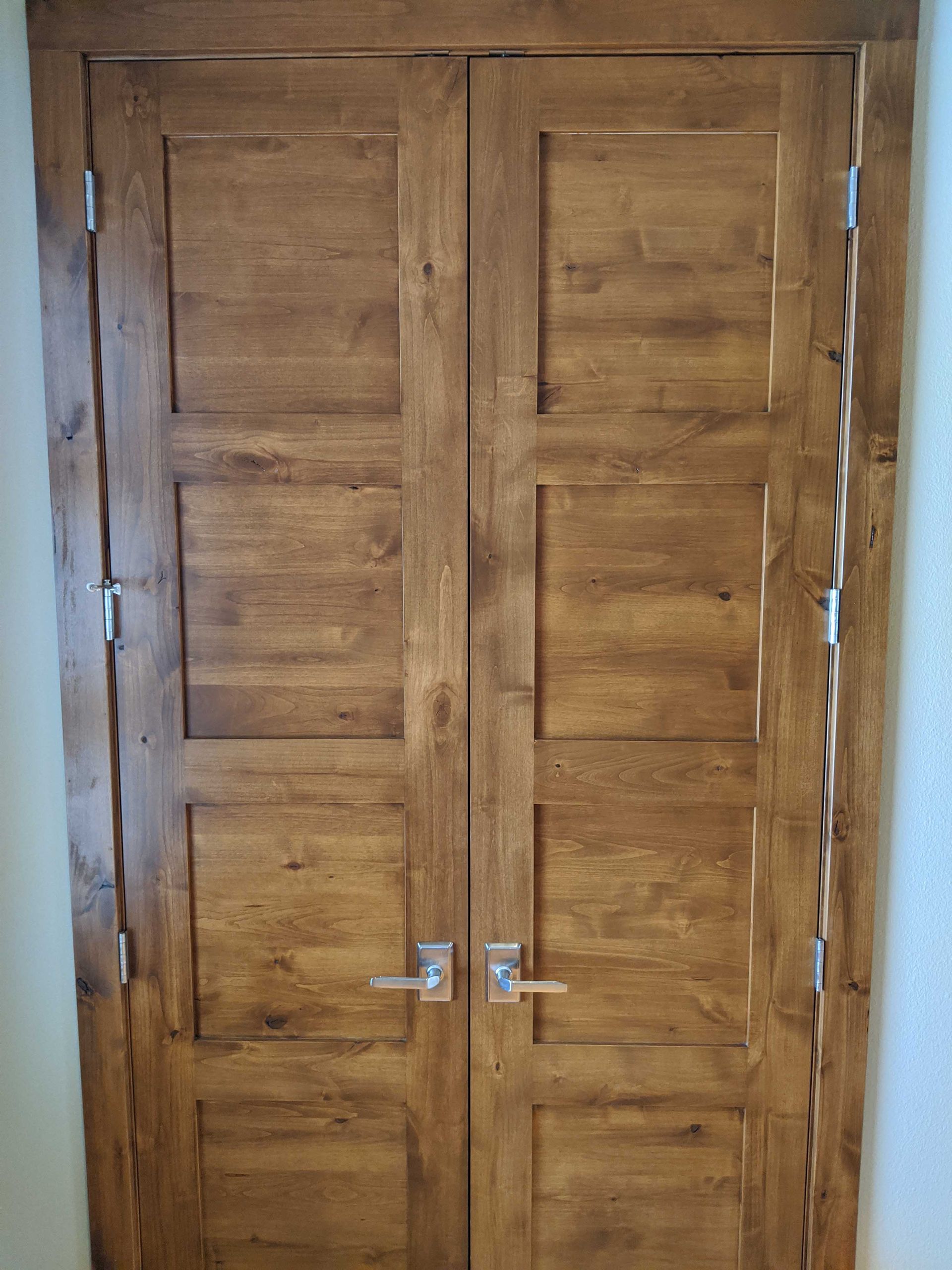 Double wood doors with a natural brown finish and rectangular paneling, featuring silver handles and hinges.