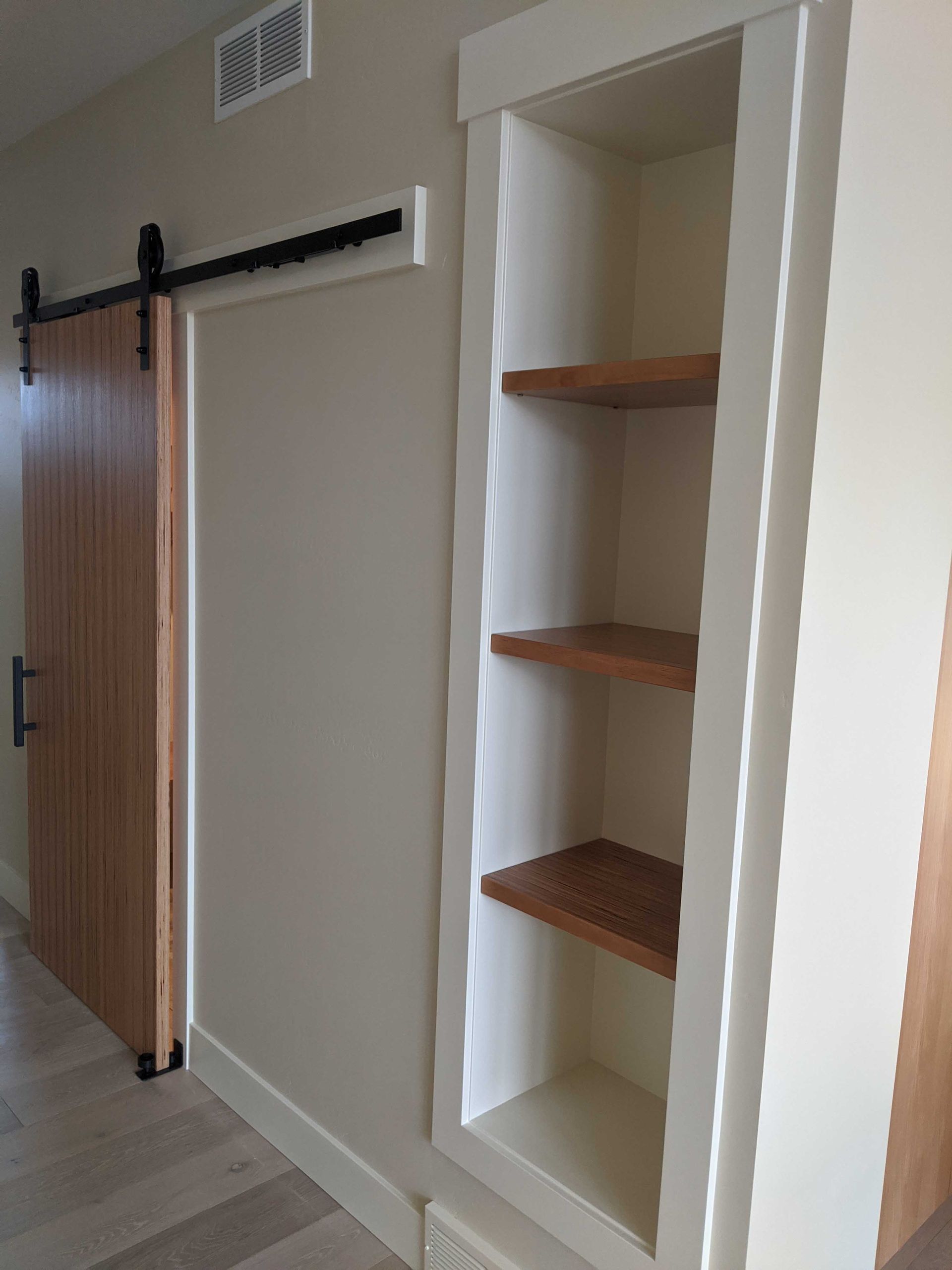 A wooden barn door on a black sliding rail next to a built-in recessed wall shelf with three wooden shelves.