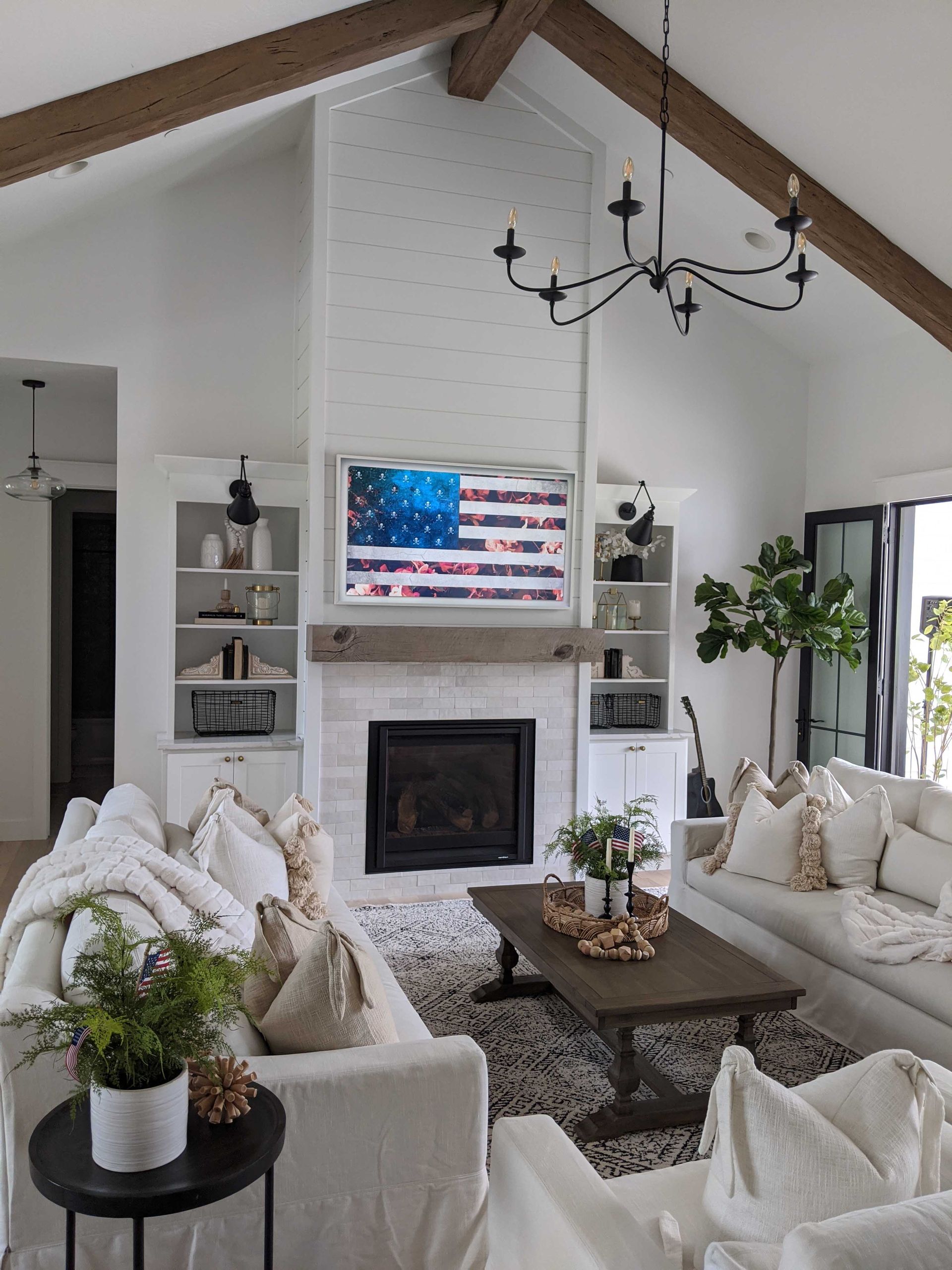 A white living room with a brick fireplace, rustic wood mantel, shiplap wall, vaulted ceiling with beams, and sofas.
