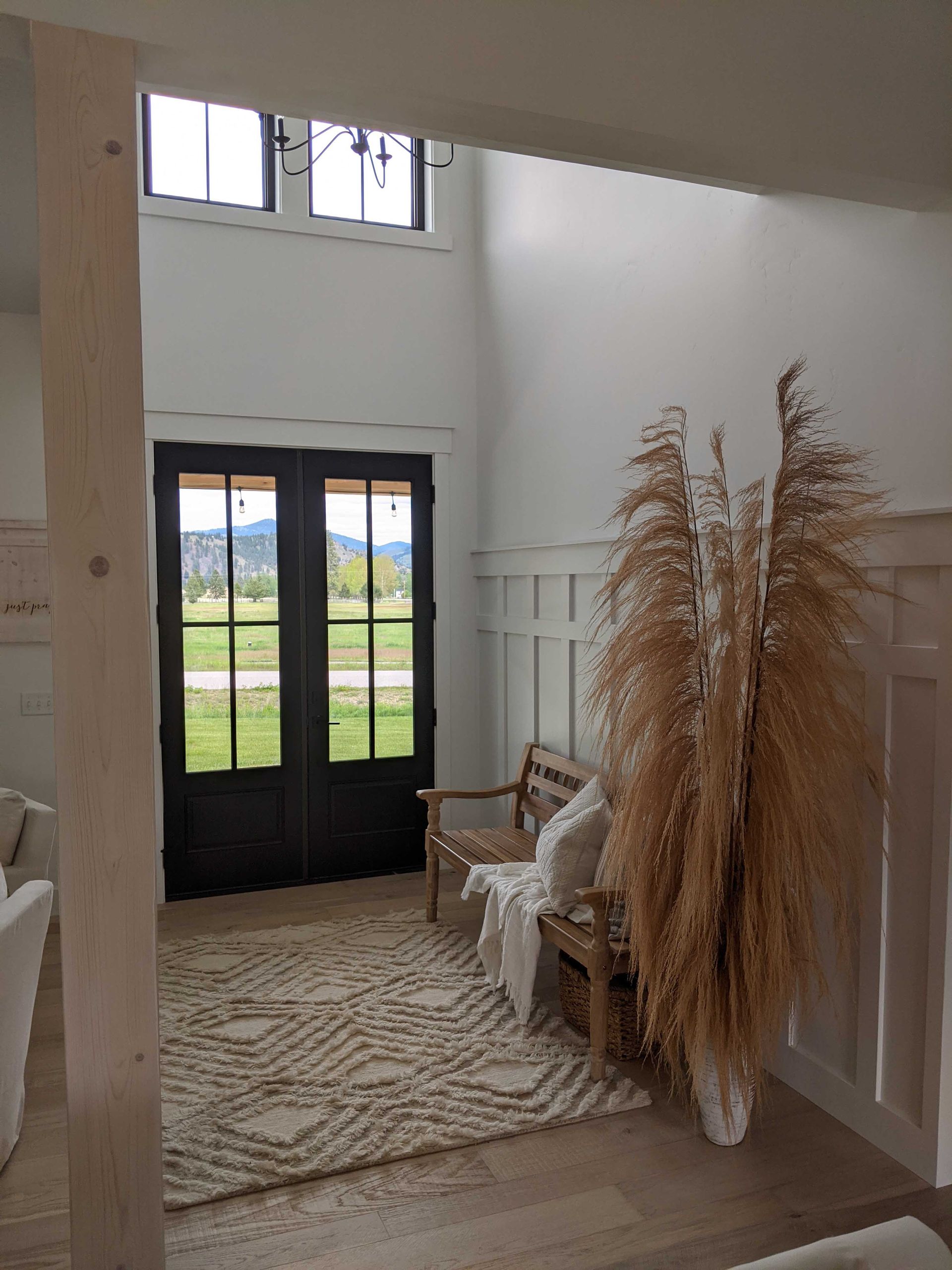 A bright entryway with a double black door, a patterned area rug, a wooden bench, and a tall pampas grass arrangement.