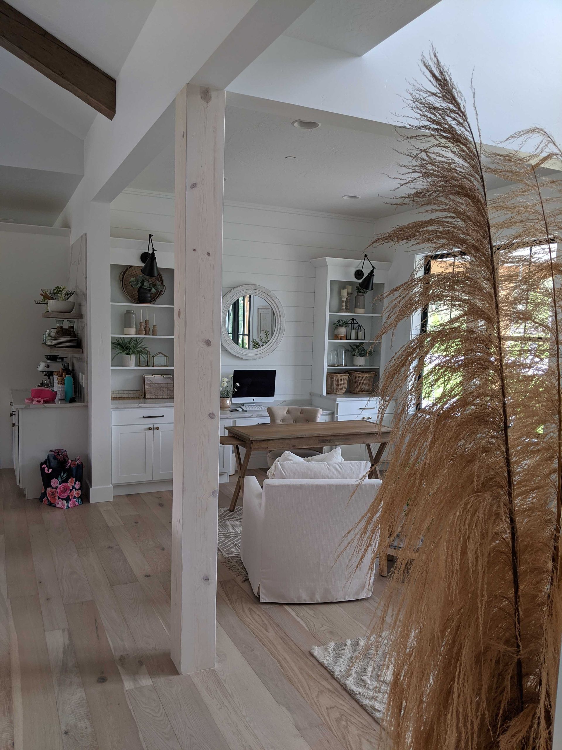 A bright, modern home office with white built-in shelves, a desk, a slipcovered chair, and a large pampas grass plant.