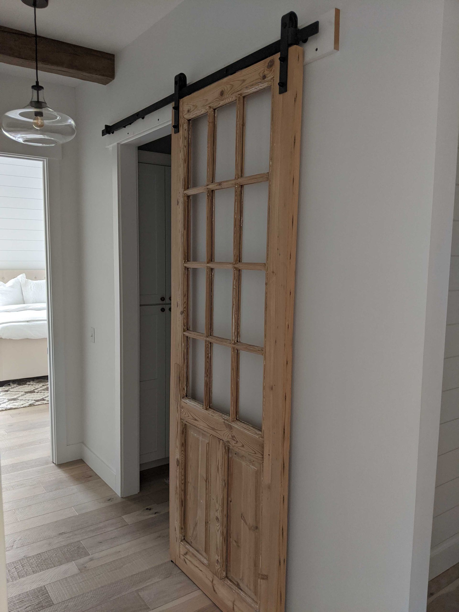 A rustic wooden barn door with glass panels hangs on a black metal sliding track against a white wall in a hallway.