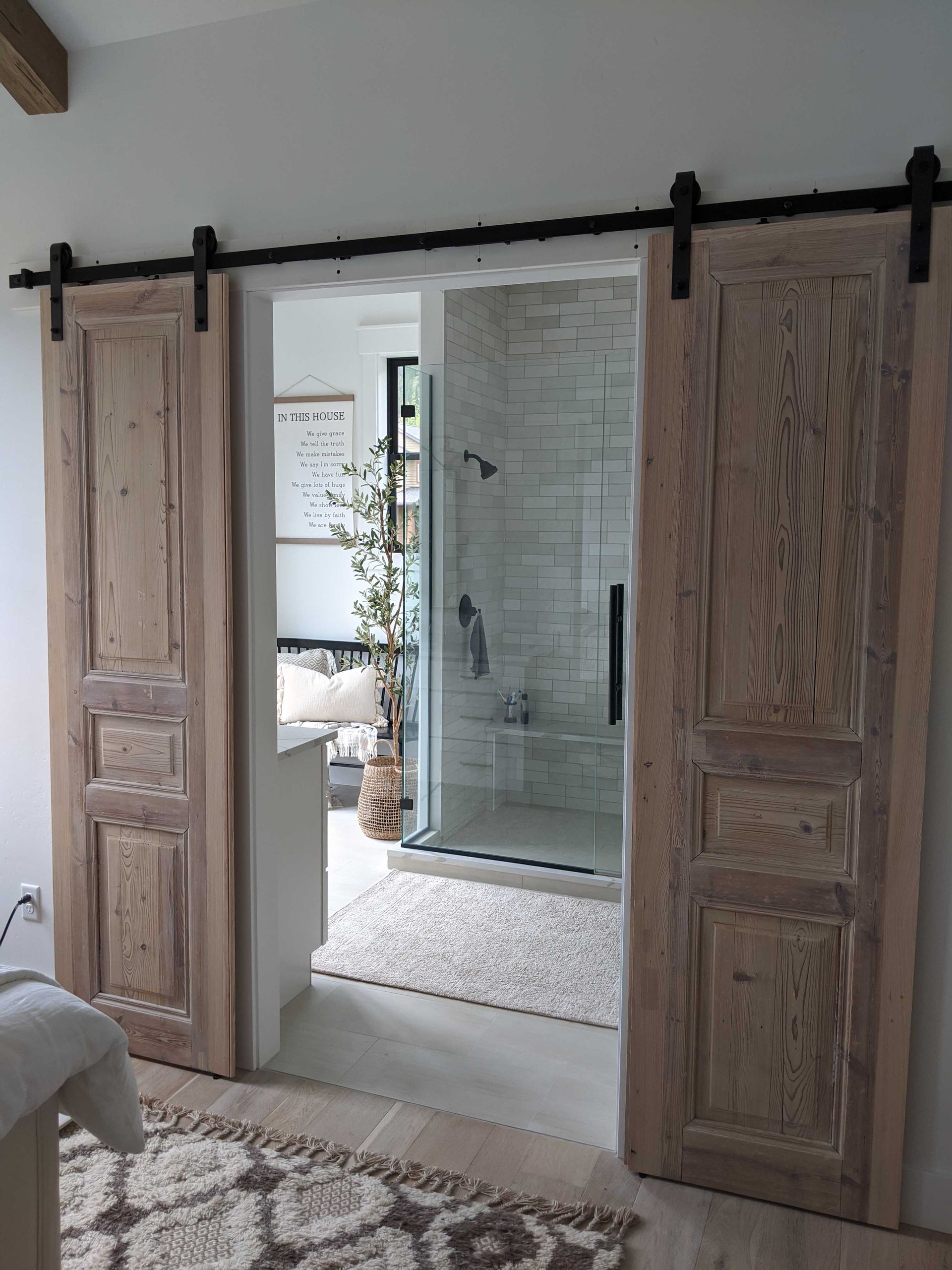 Two natural wood barn doors slide on a black metal track, revealing a bright, modern bathroom with a glass shower.